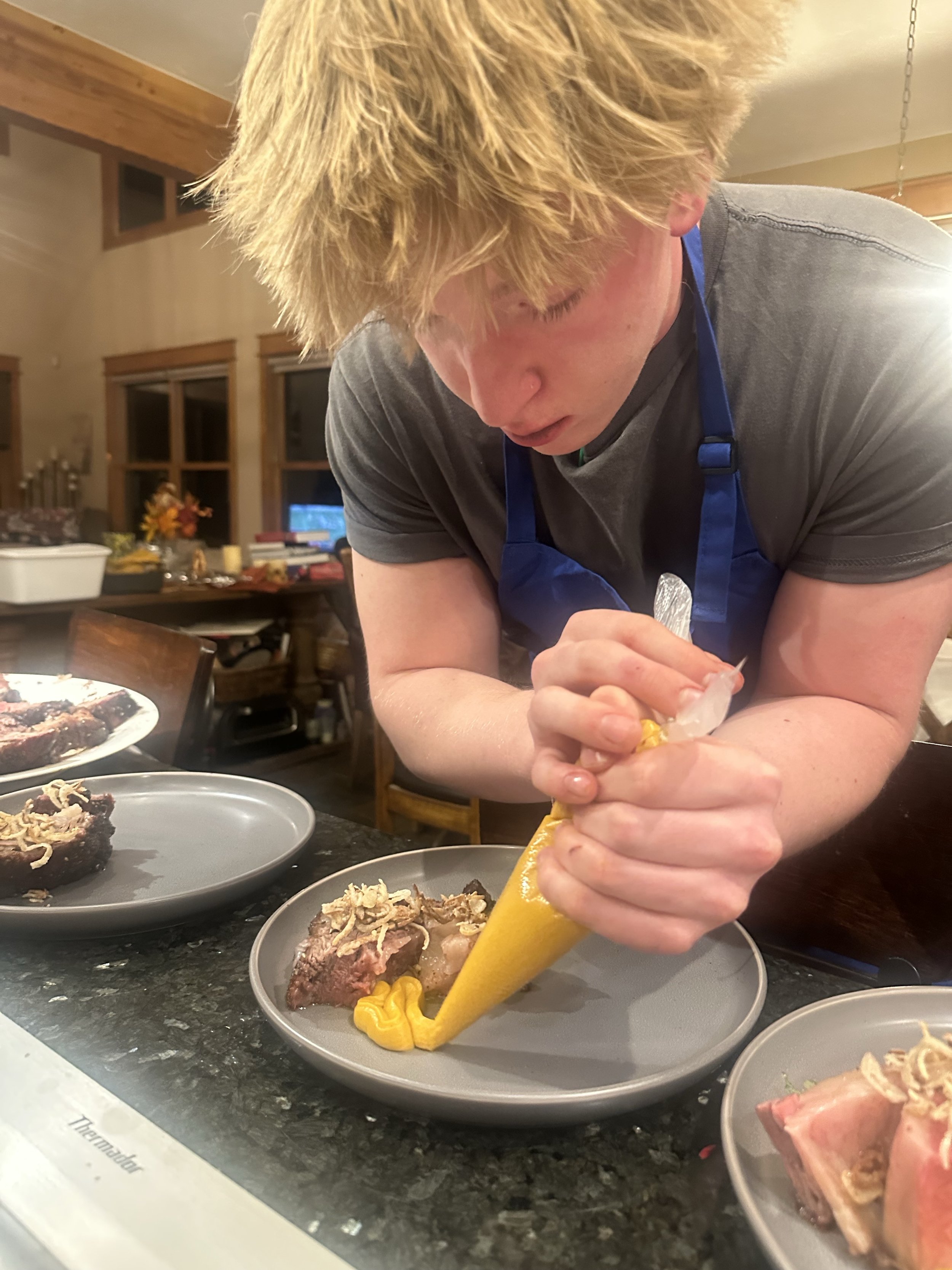 A person with blonde hair wearing a gray shirt and a blue apron is piping yellow sauce onto a plate of cooked meat with garnishes, in a warmly lit kitchen or dining area.