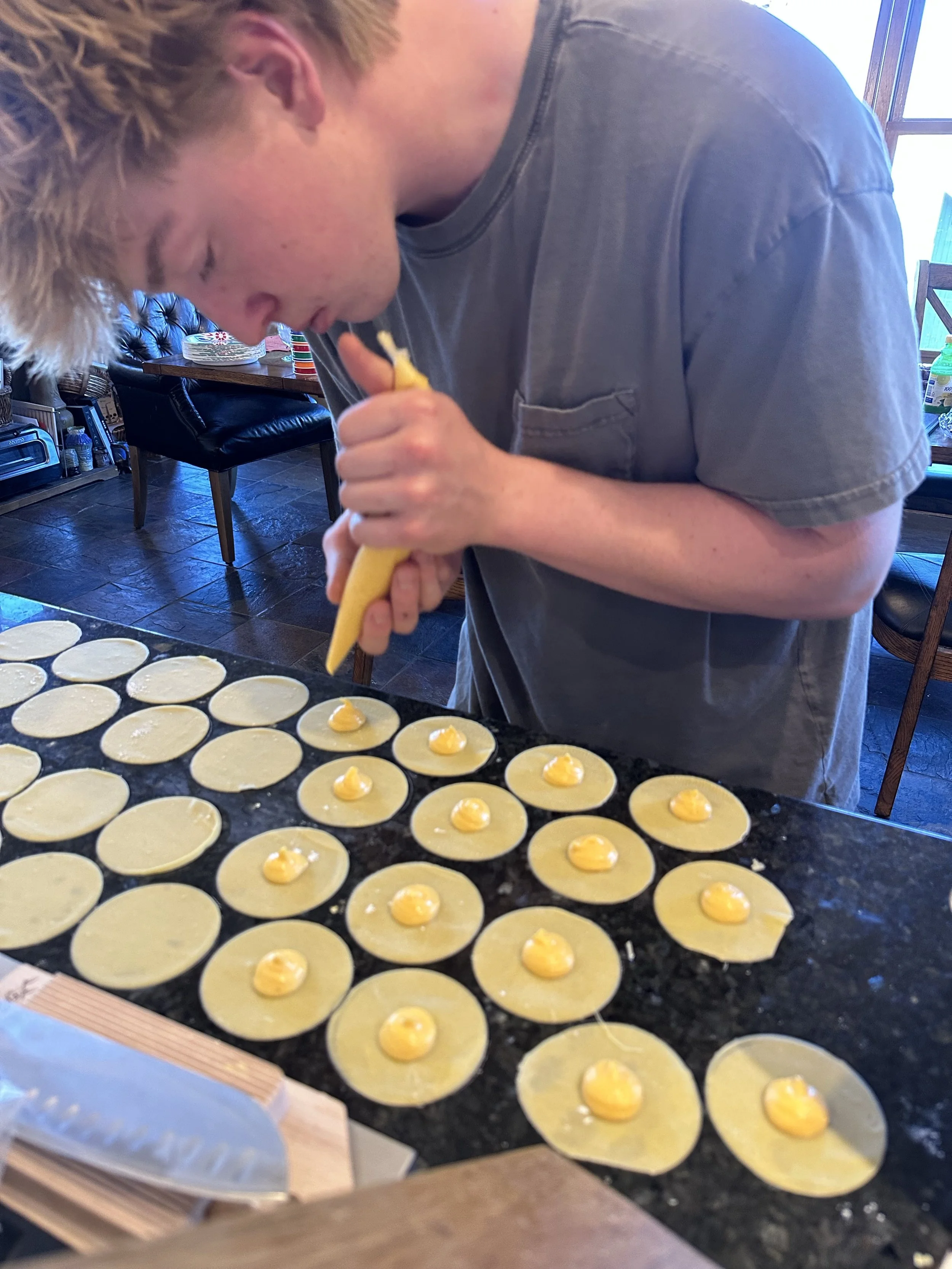 A person in a gray t-shirt is piping yellow filling onto circular pastry wrappers on a black countertop in a kitchen.