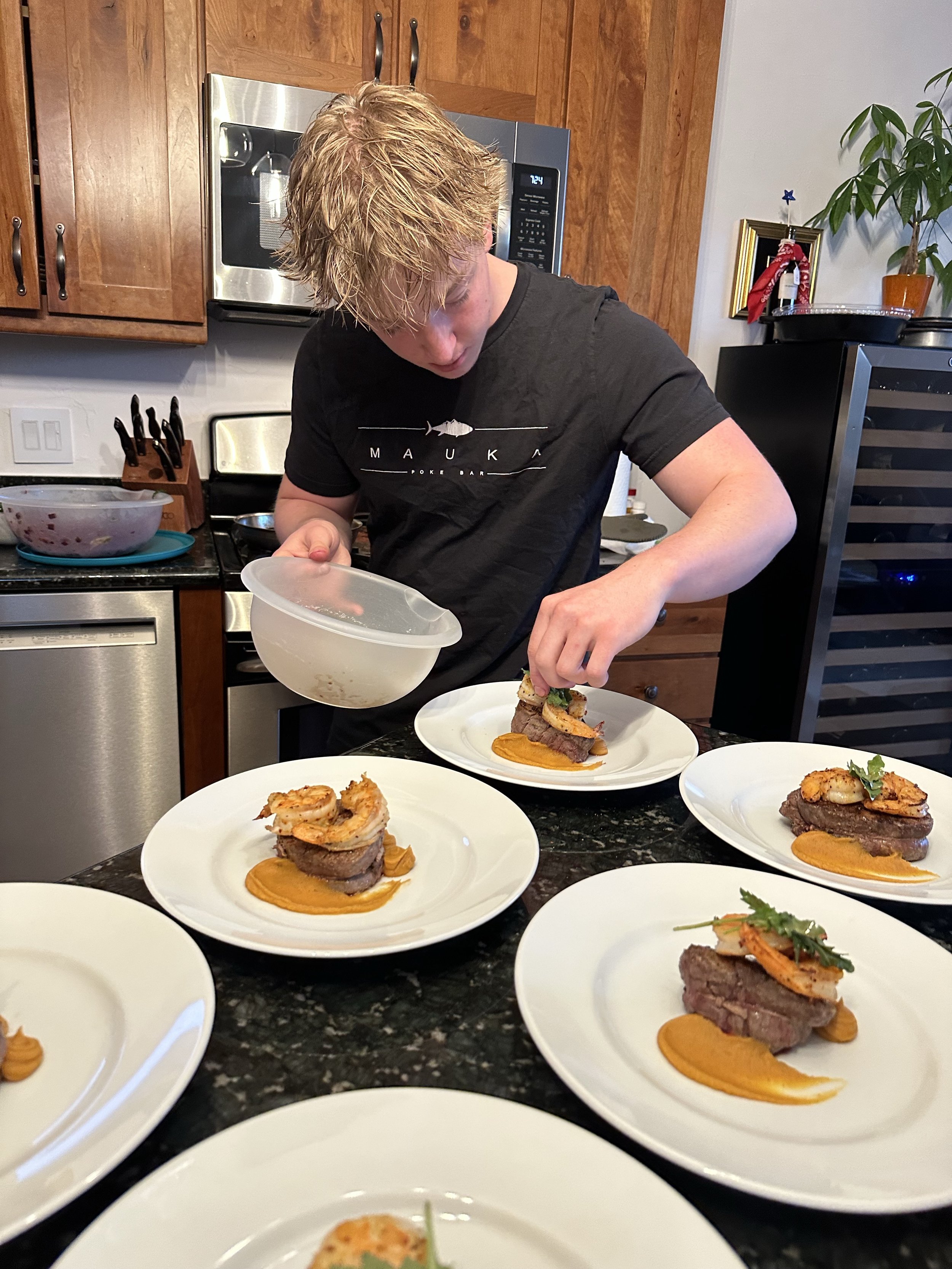 A person with blond hair in a black t-shirt preparing dishes with steak, shrimp, and sauce on a kitchen counter with several plated servings.