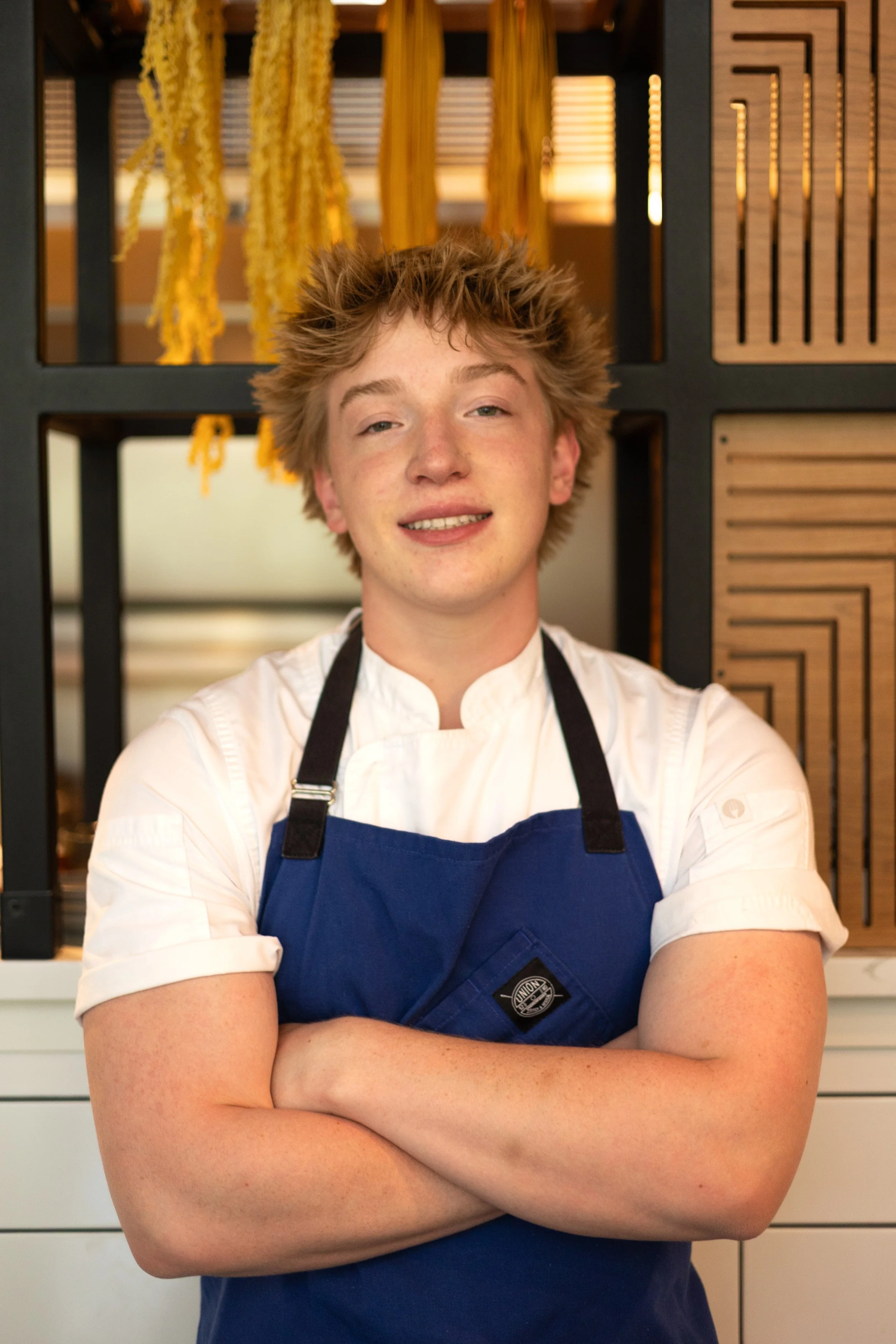 A young male chef stands with his arms crossed in a modern kitchen, wearing a white chef's coat and a blue apron.