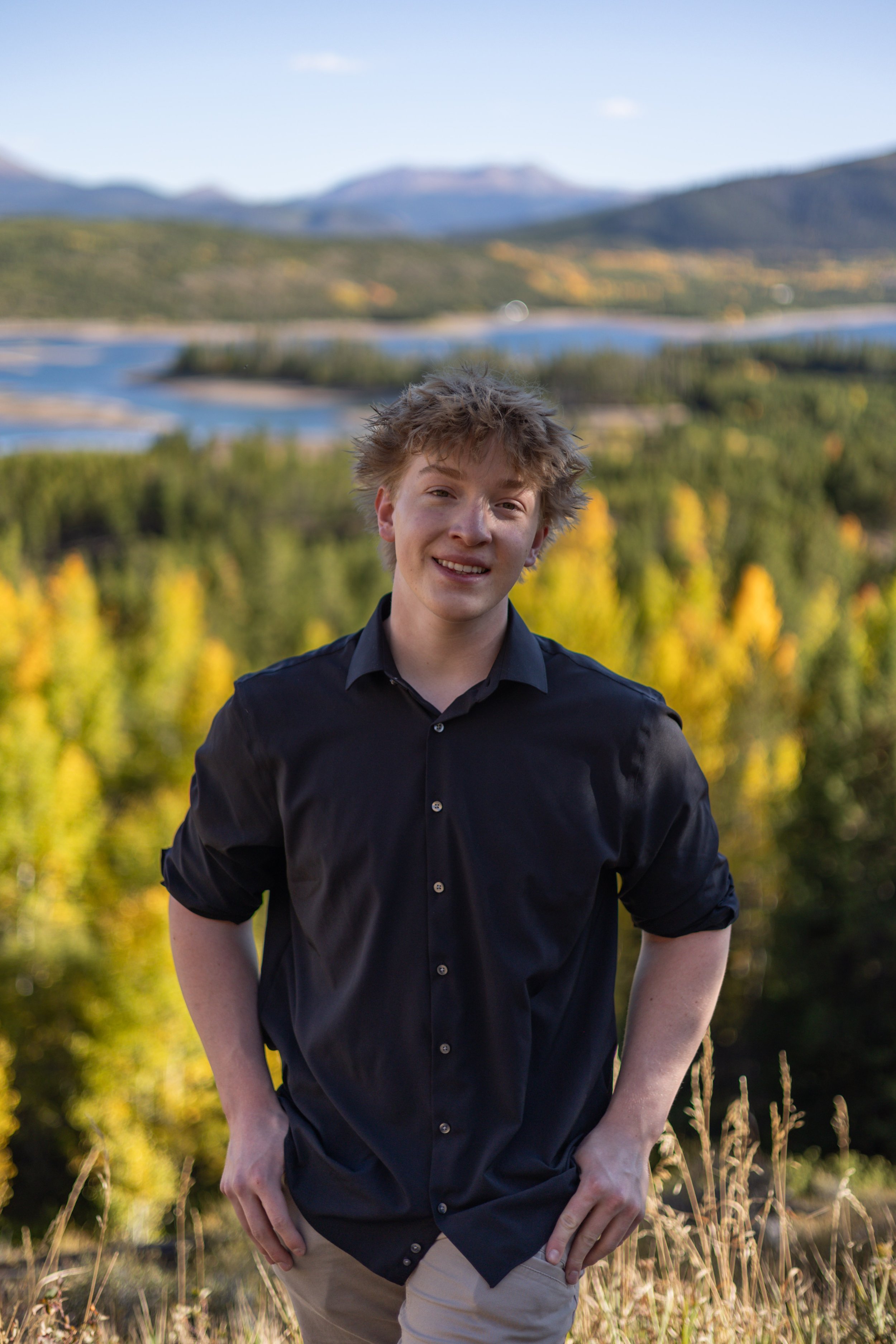 Young man with curly hair smiling outdoors with a scenic landscape of trees, a lake, and mountains in the background.