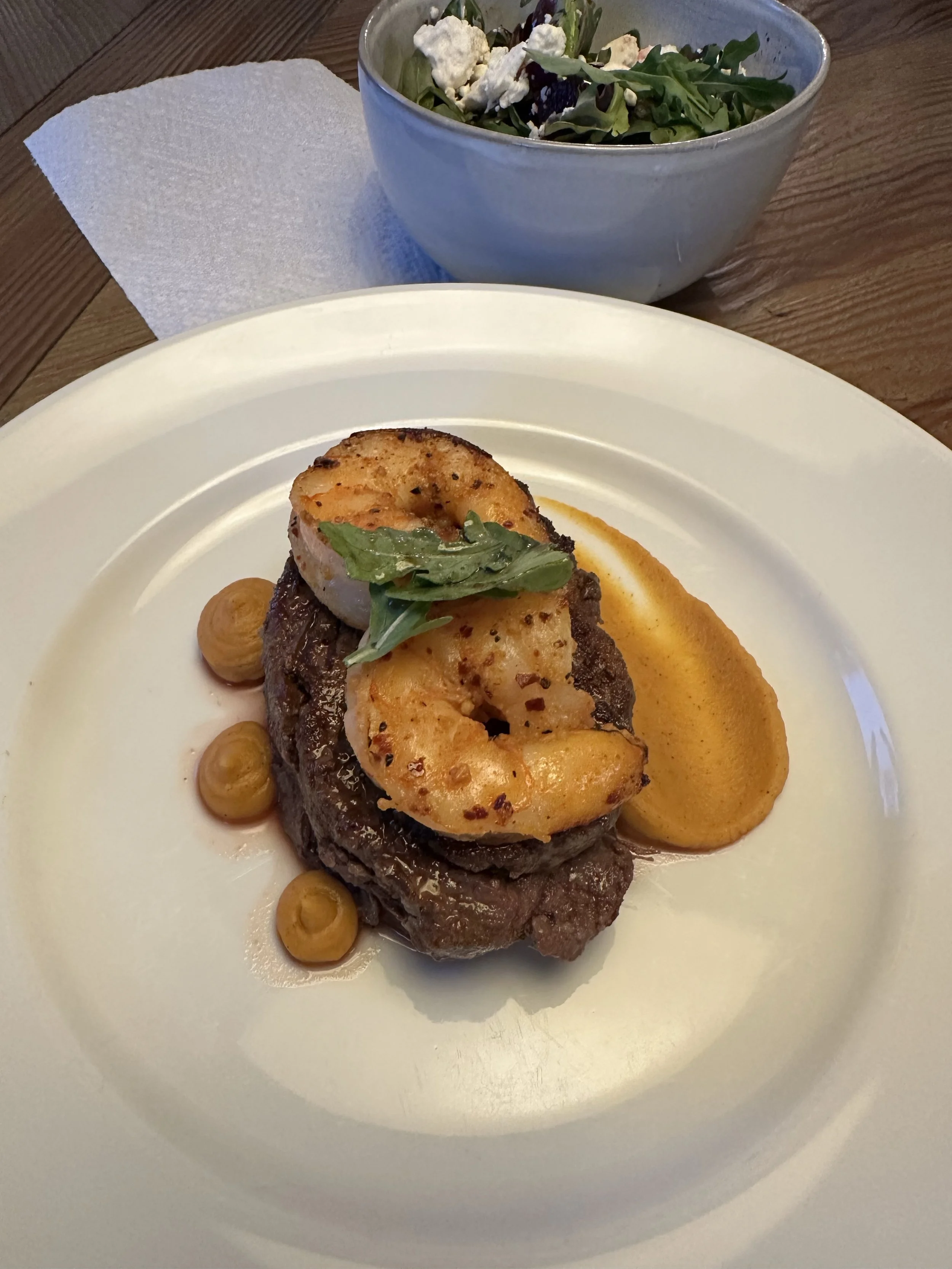 A gourmet plated dish featuring a piece of grilled steak topped with seared shrimp and garnished with a small green herb leaf, accompanied by a smear of orange sauce and small dollops of a yellowish puree on a white plate. 