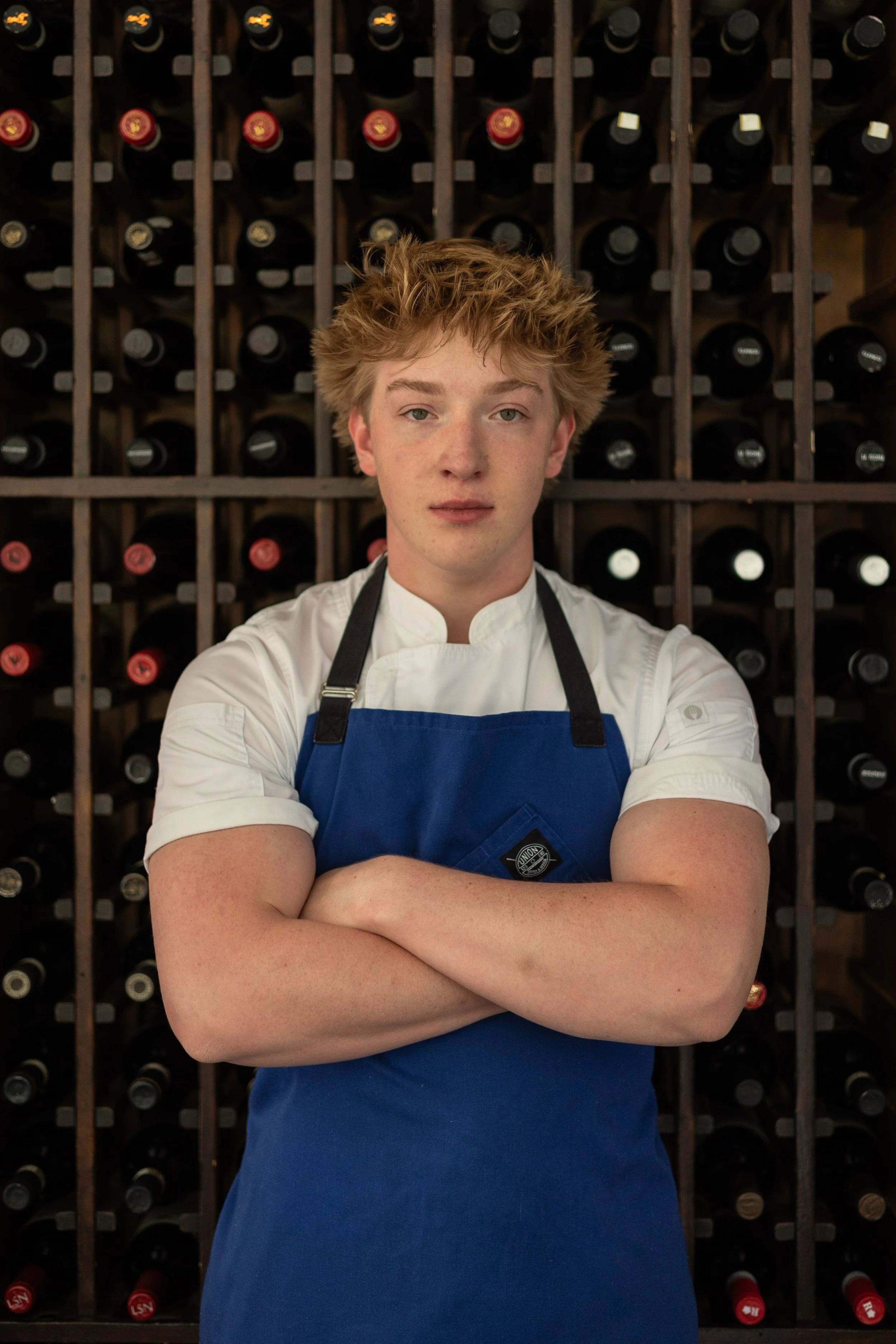 Curtis Hammer staring into the camera with his arms crossed in an apron. Wine bottles in the background.