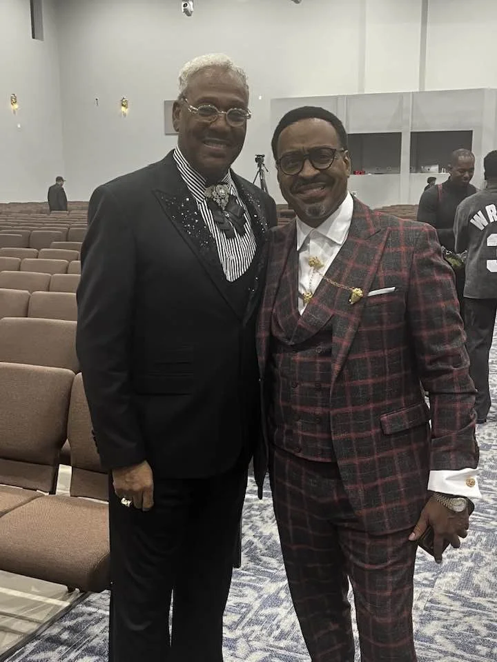 Two men in suits standing in an elevator or conference room with empty chairs behind them.