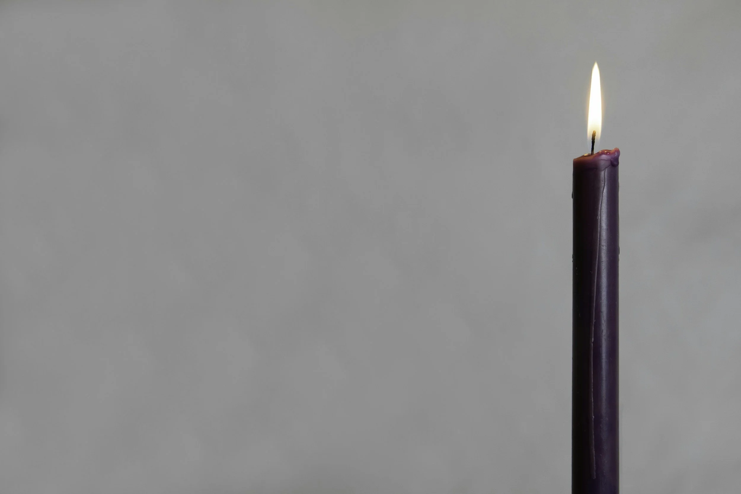 A purple candle burning with a small flame on a plain gray background.