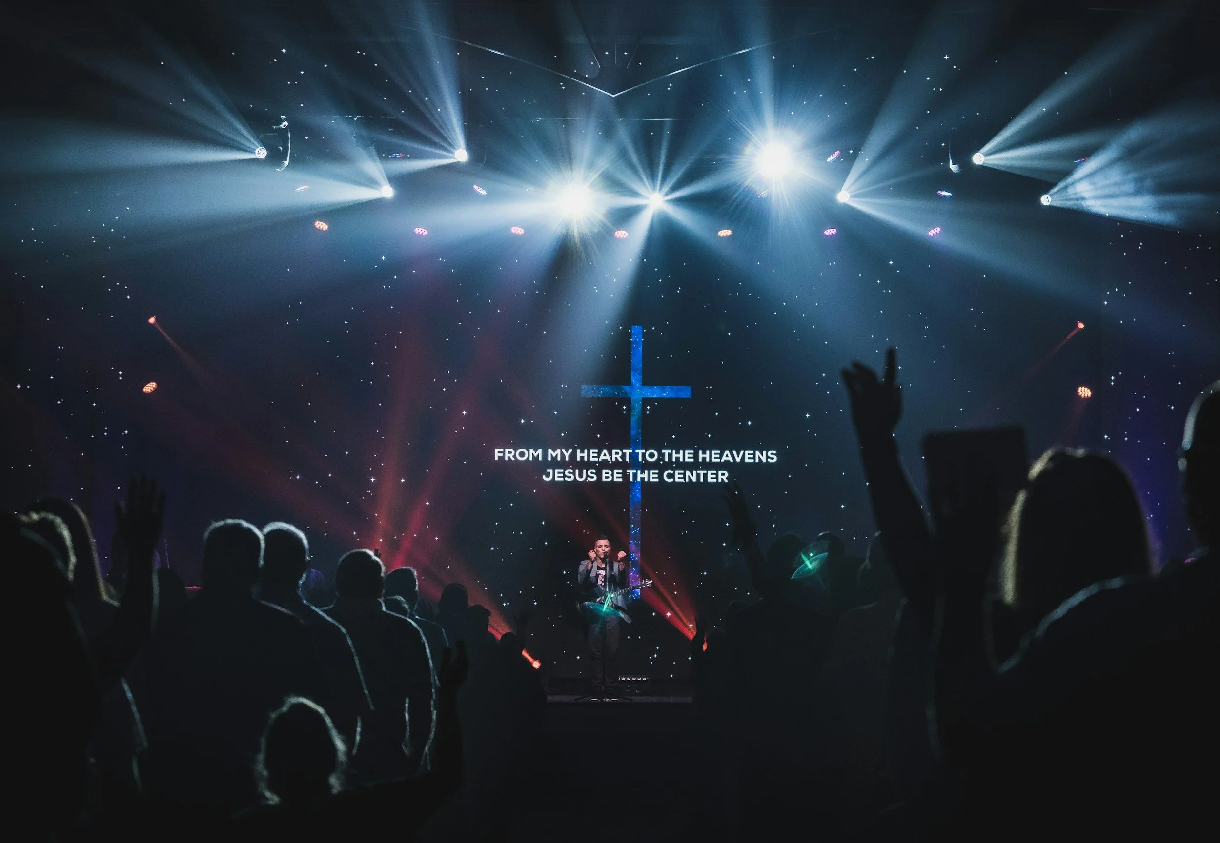 Crowd at a church service with a worship leader on stage, silhouetted against a starry background with bright lights and a large cross, displaying lyrics: 'FROM MY HEART TO THE HEAVENS JESUS BE THE CENTER.'