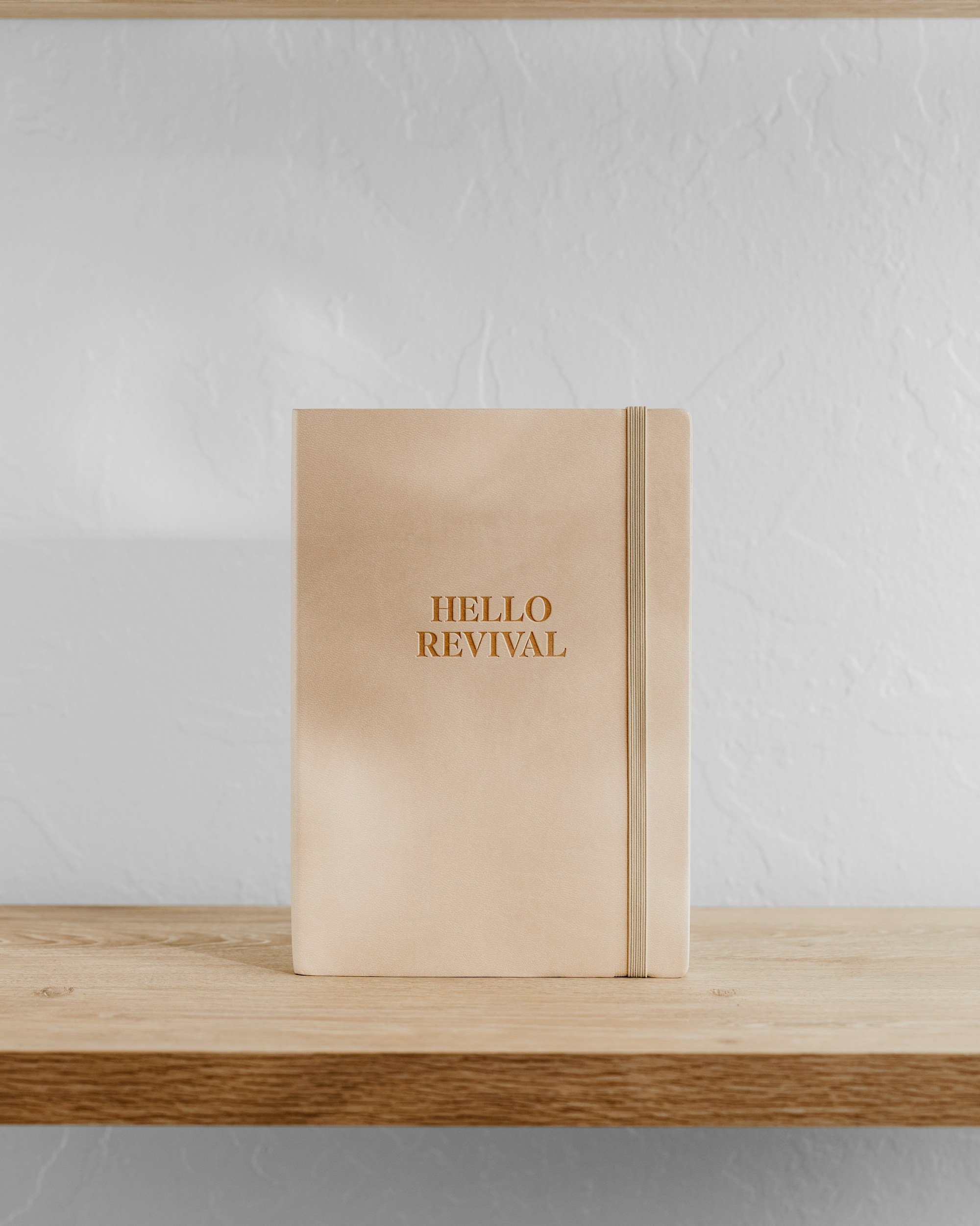 Beige notebook with gold embossed text reading "HELLO REVIVAL" and a beige elastic band, placed on a wooden surface against a white textured wall.