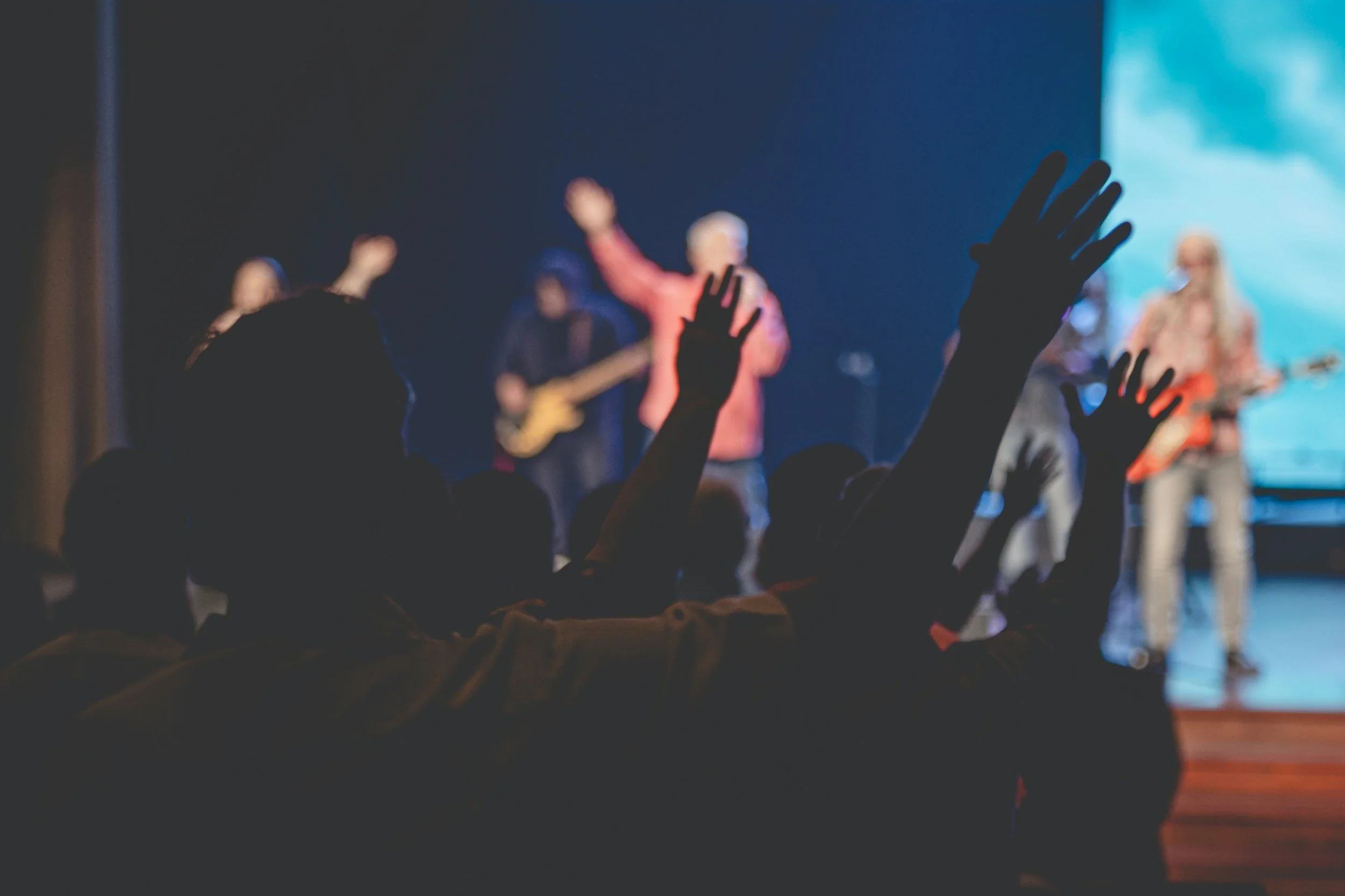 Audience members raising their hands during a live music performance on stage with band members playing instruments.