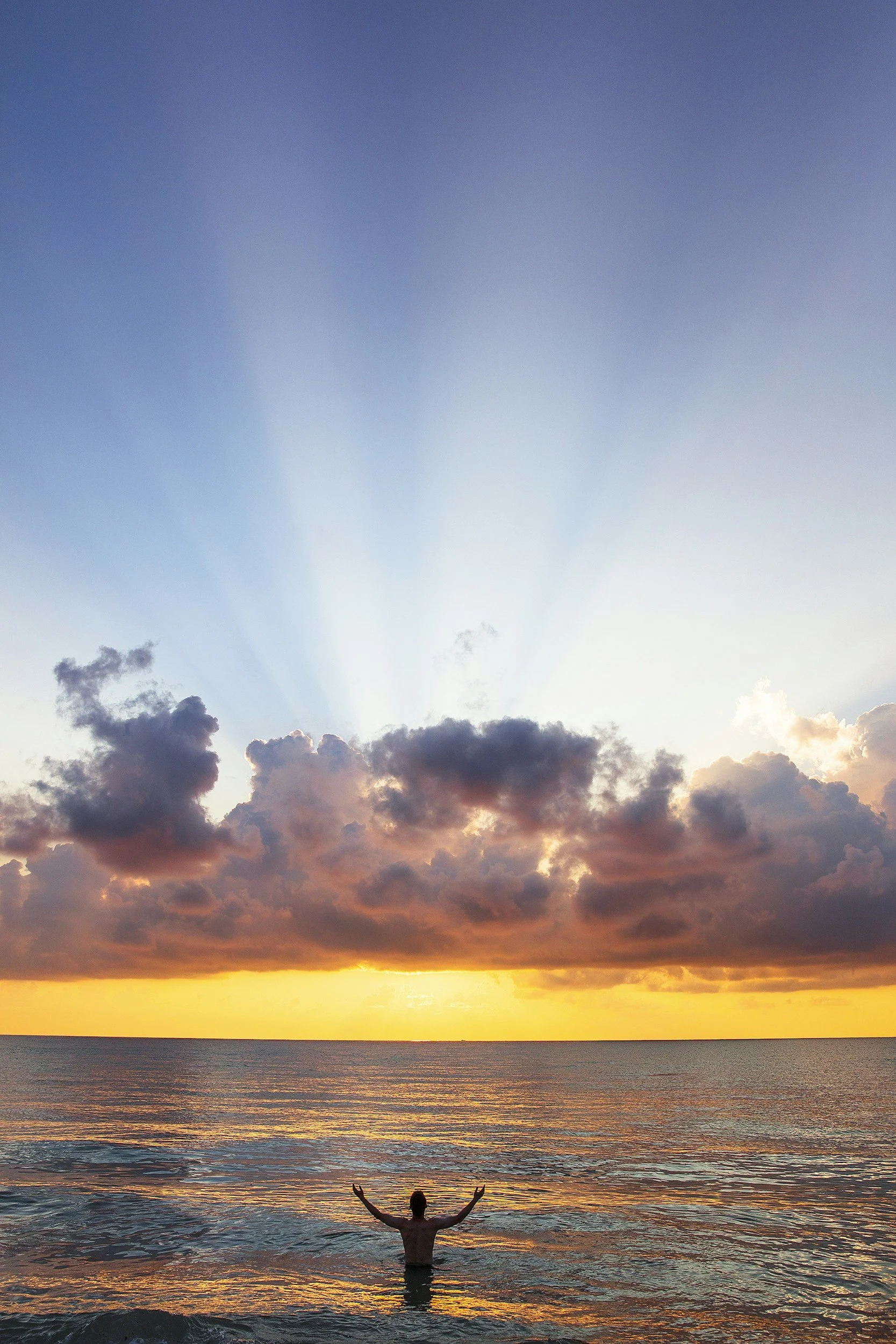 Person standing in the ocean with raised arms at sunset, facing the sky with rays of sunlight breaking through clouds.