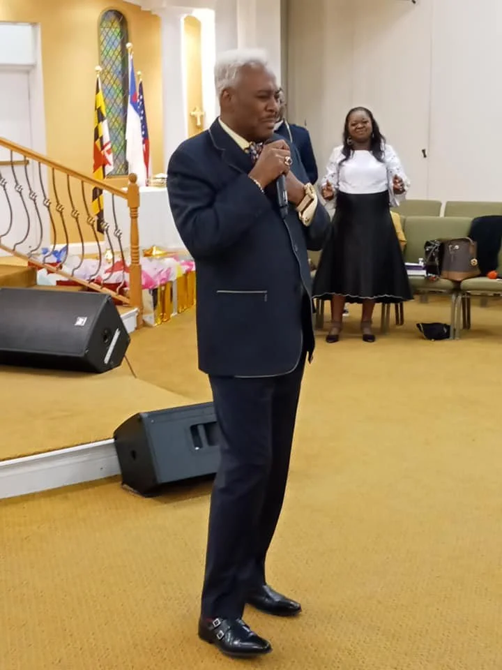 A man with gray hair dressed in a dark suit, holding a microphone, standing in a church or event hall. A woman in a white blouse and black skirt is in the background, smiling, near a row of chairs and flags.