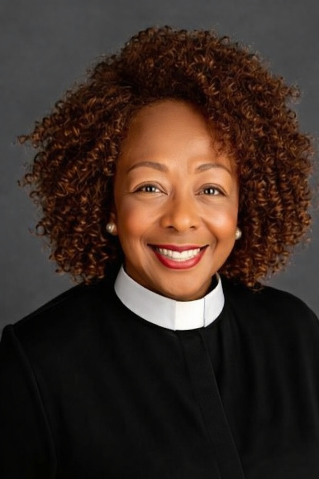 Dr. Deborah Wortham, with curly reddish-brown hair, wearing a black clergy shirt with a white clerical collar, pearl earrings, and red lipstick, against a dark gray background.