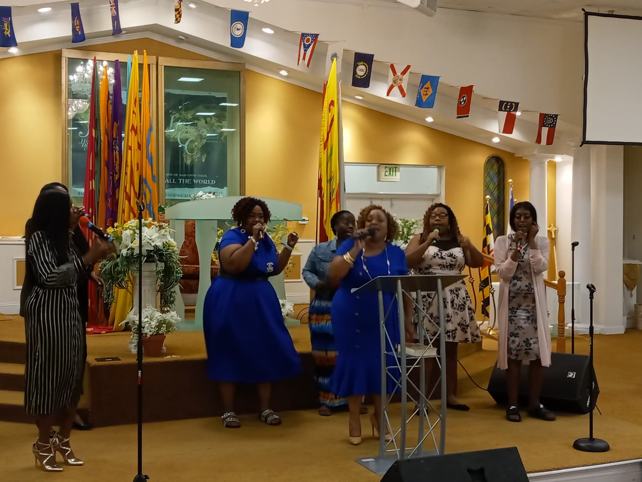 Five women singing on a church stage with flags and floral arrangements in the background.