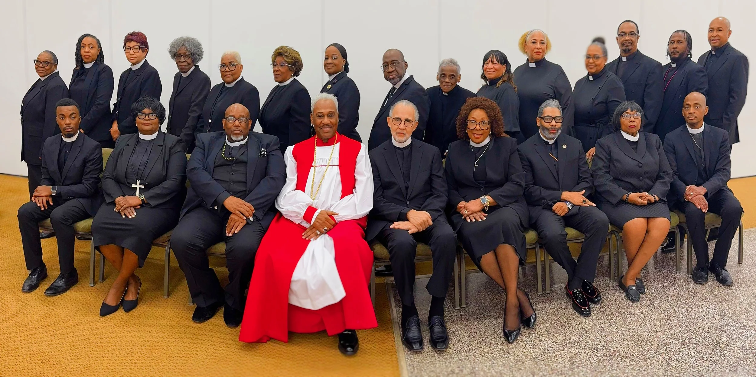 Group photo of religious leaders dressed in black and clerical attire, with one person in a white and red robe, sitting and standing in a room with beige carpet and plain white walls.