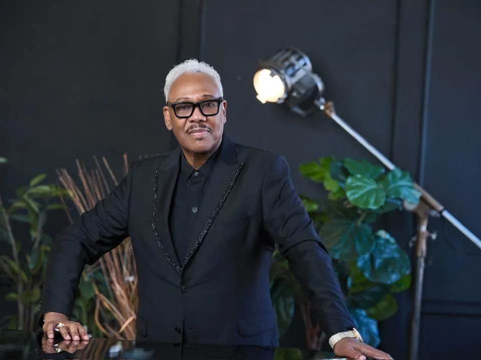 A man with gray hair and glasses wearing a black suit and shirt, standing in a studio with a black background, a large studio light, and green plants.