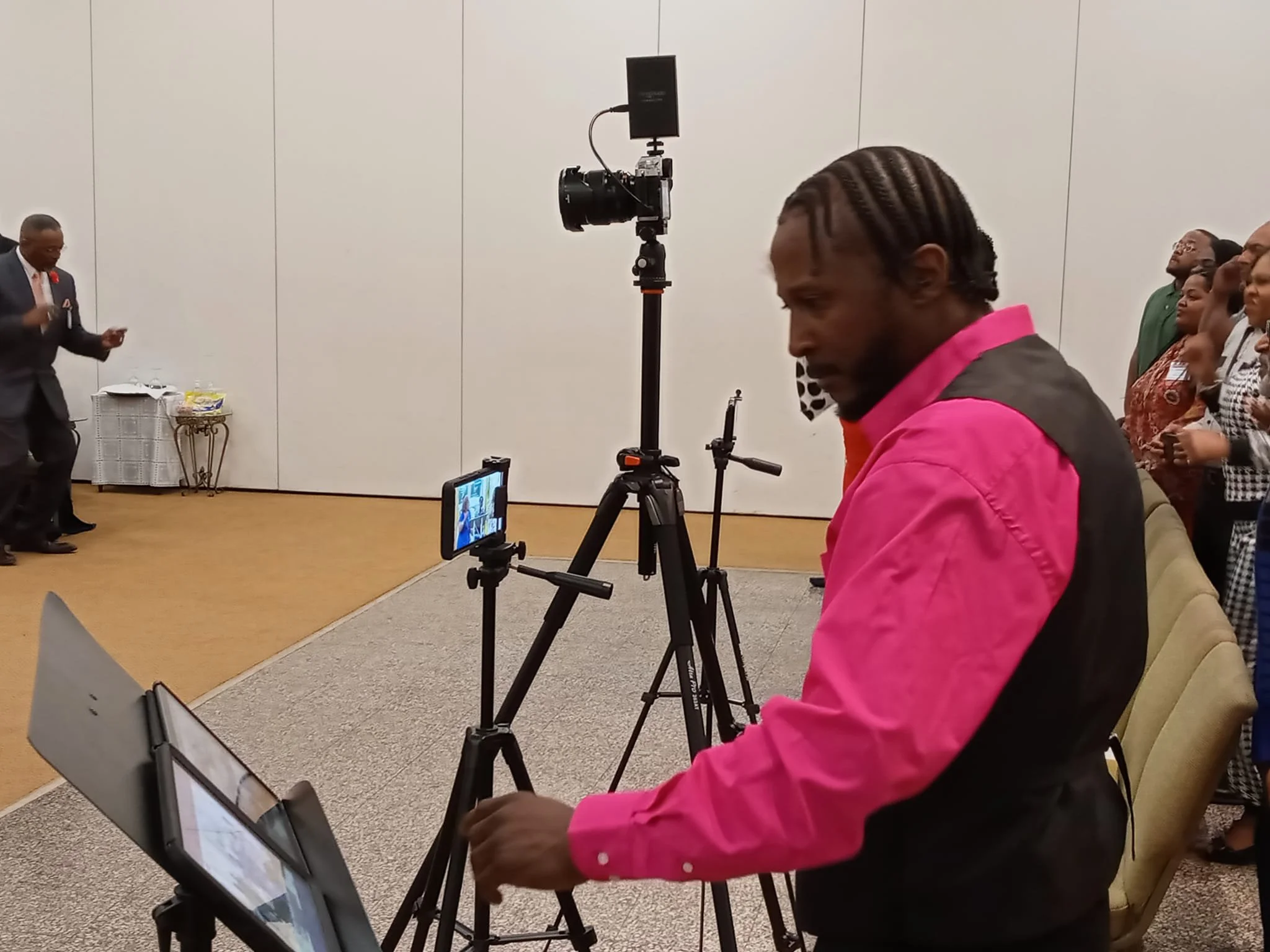 A man in a pink and black shirt operating a video camera on a tripod in a room with people standing and dancing in the background.