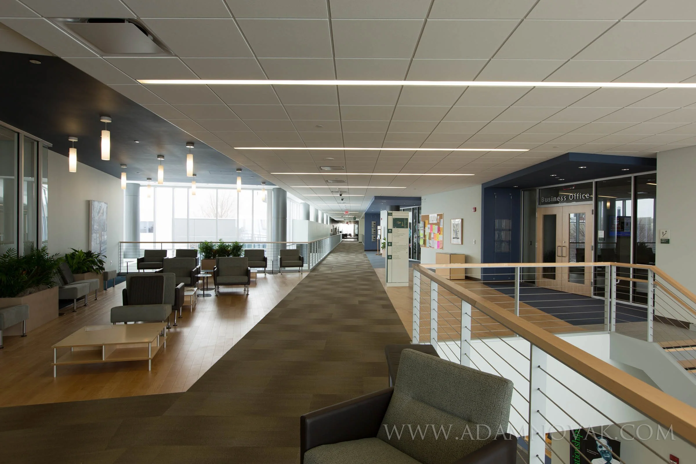 The image shows a modern, spacious interior of an office or public building with seating areas, large windows letting in natural light, and a hallway leading to a business office entrance.