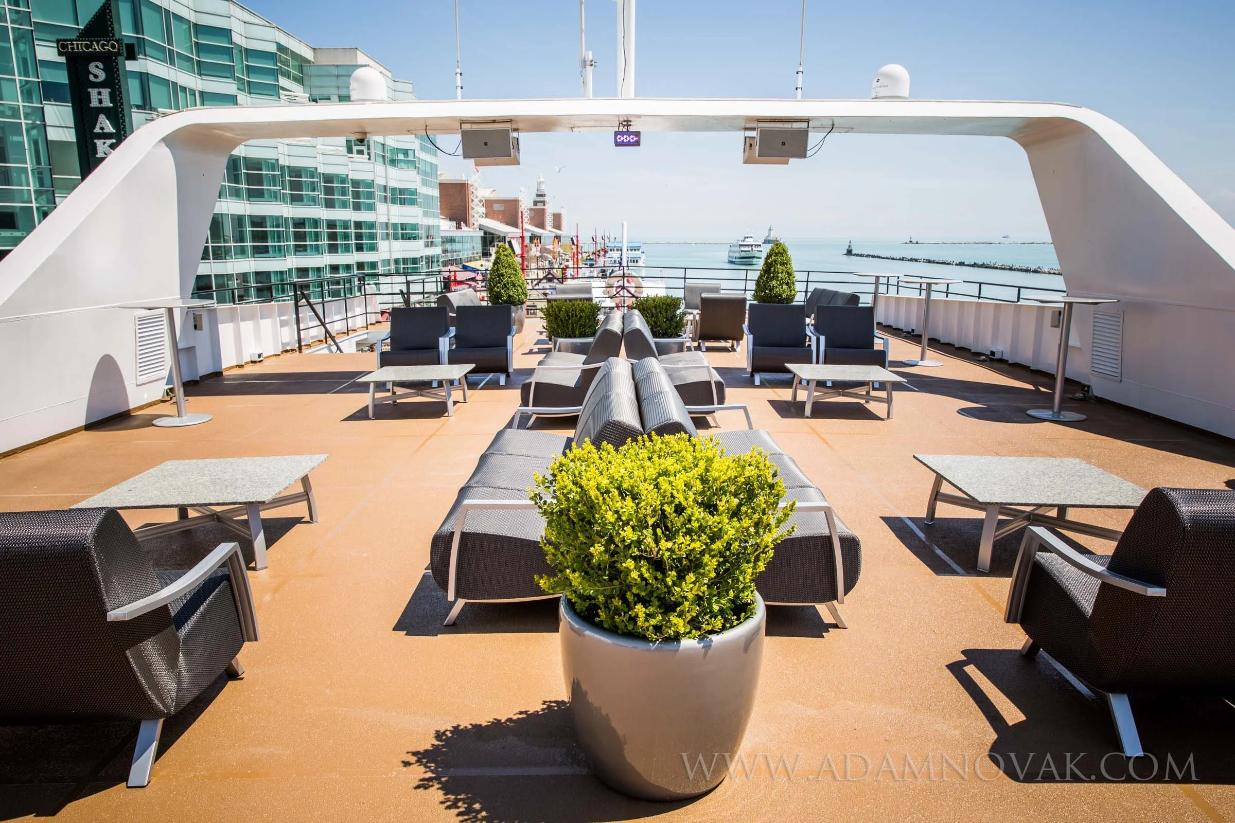 Empty outdoor deck with lounge chairs, small tables, and potted plants, overlooking a waterway and boats with city buildings in the background.