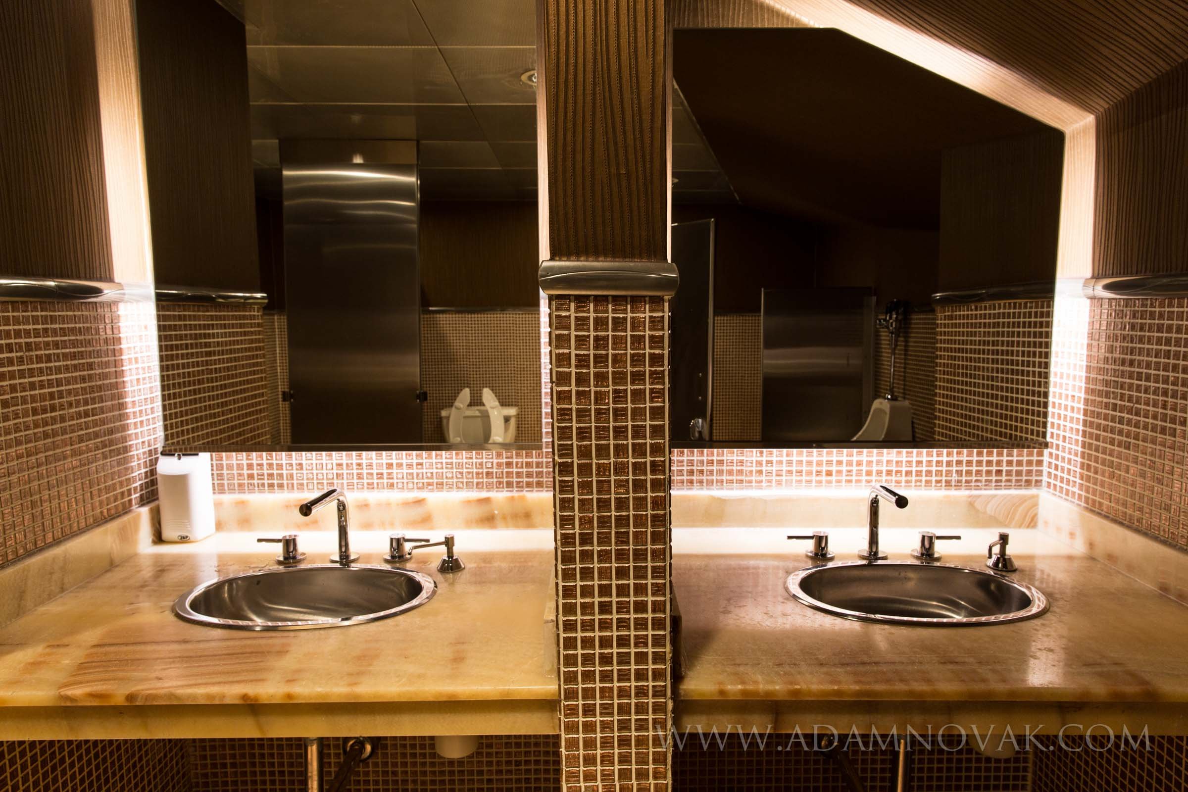 Public restroom with two sinks, large mirror, brown tiled walls, and a divider in the center.