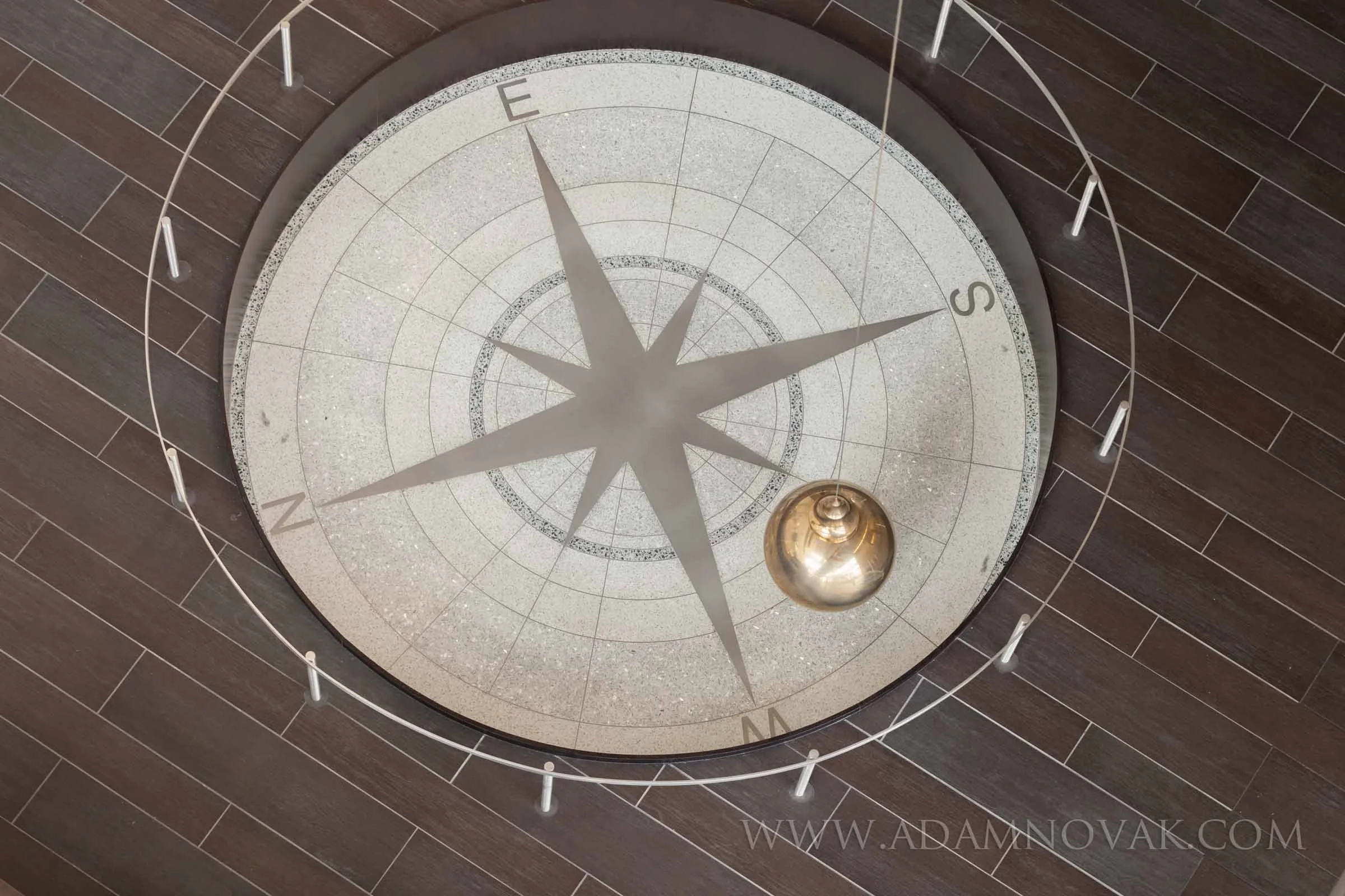 A decorative indoor floor compass with a metallic sphere in the center, surrounded by a metal guardrail, on dark wood plank flooring.