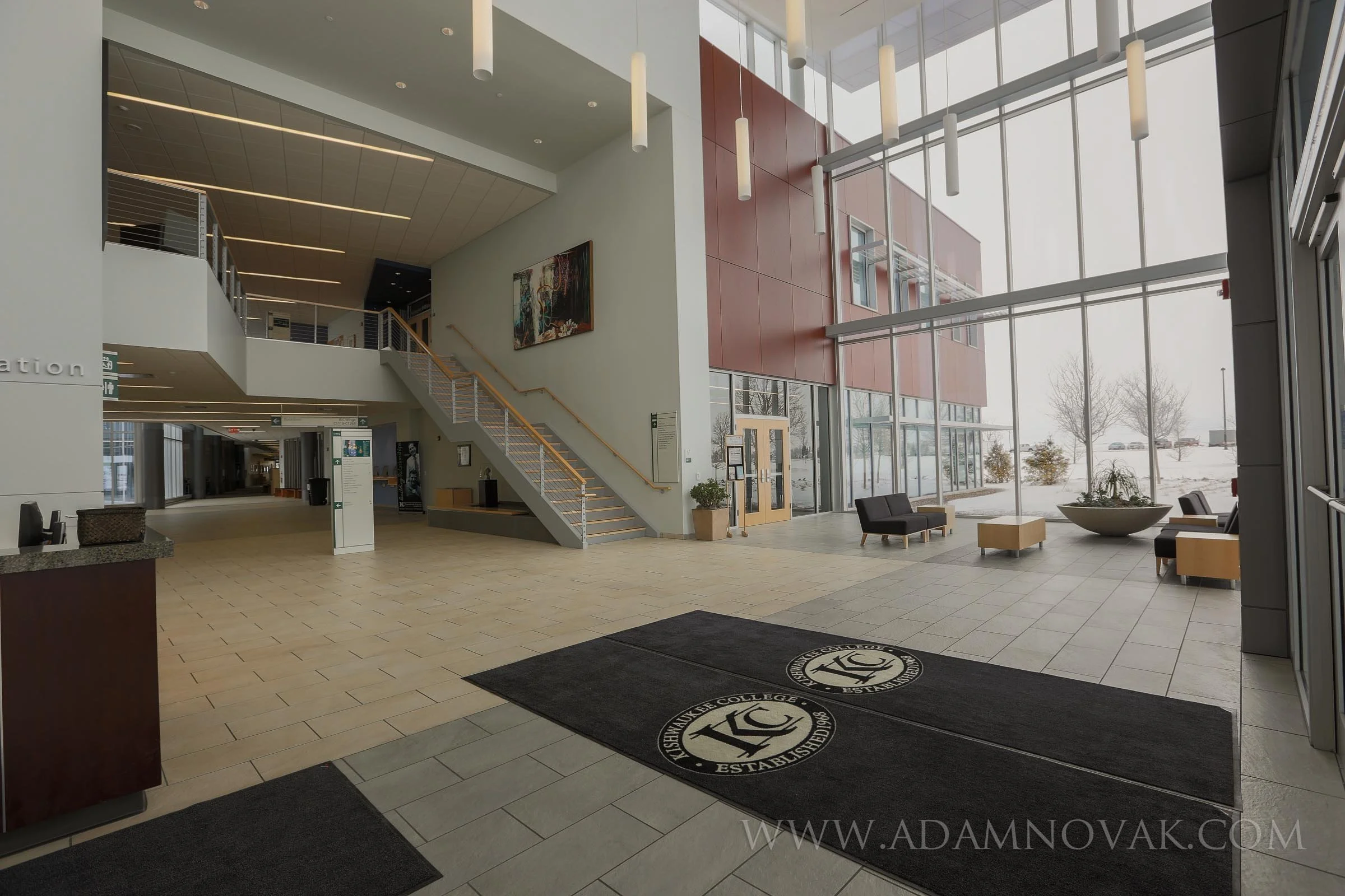 Interior of a modern college building lobby with large windows, seating area, staircase, and a rug with university logo