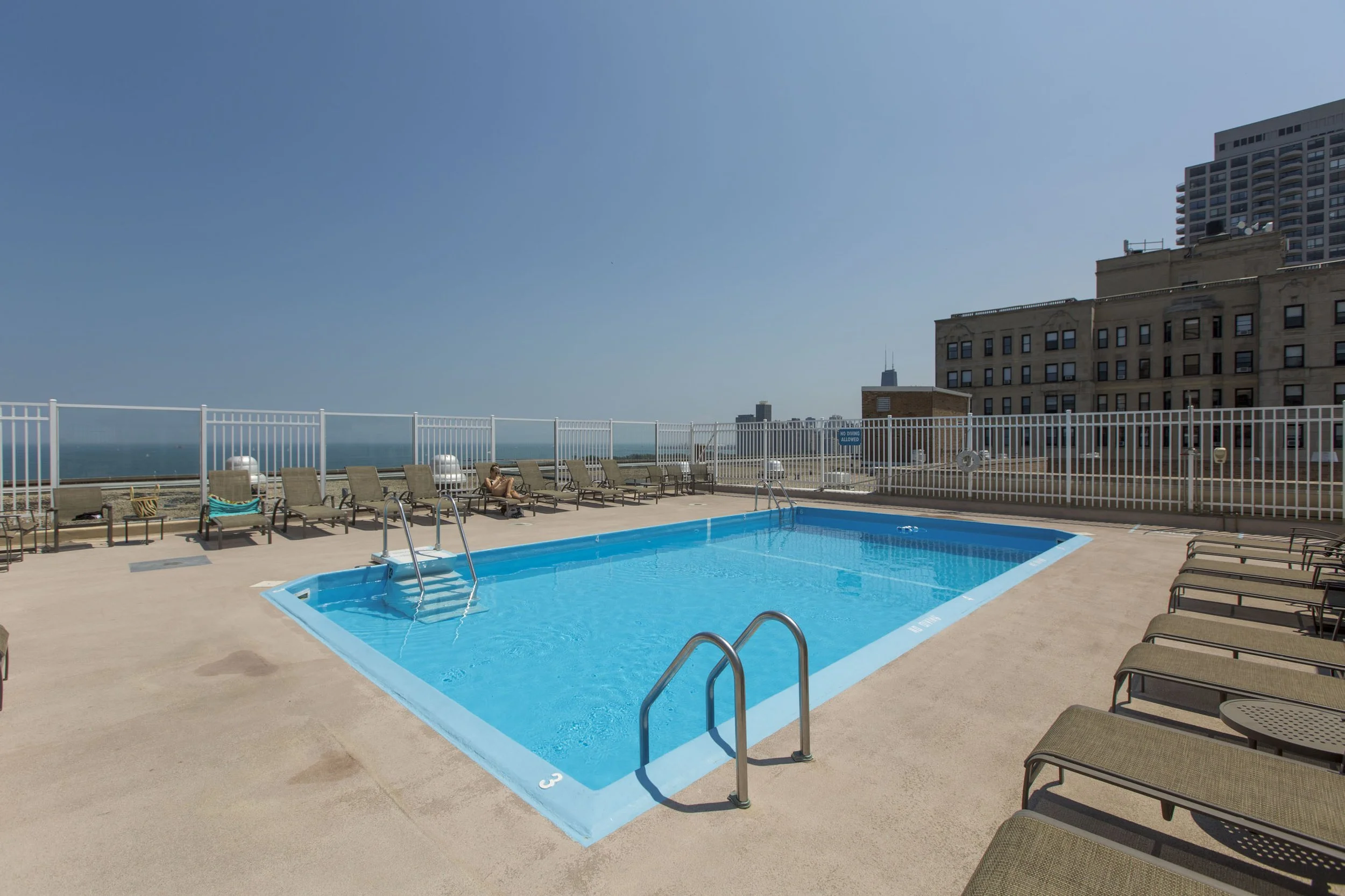 Empty rooftop swimming pool with blue water, surrounded by lounge chairs and a white safety fence, with a clear sky and city buildings in the background.