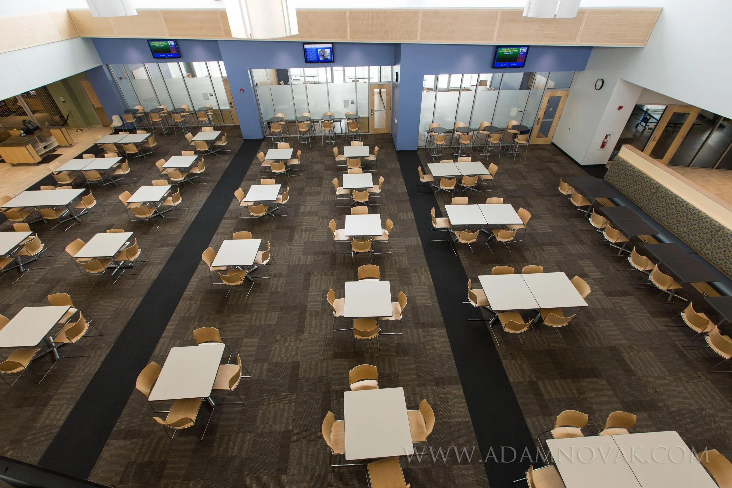 Empty modern cafeteria or dining hall with multiple square tables and beige chairs, featuring a dark patterned carpet, glass-walled private areas, and digital screens hanging from the ceiling.