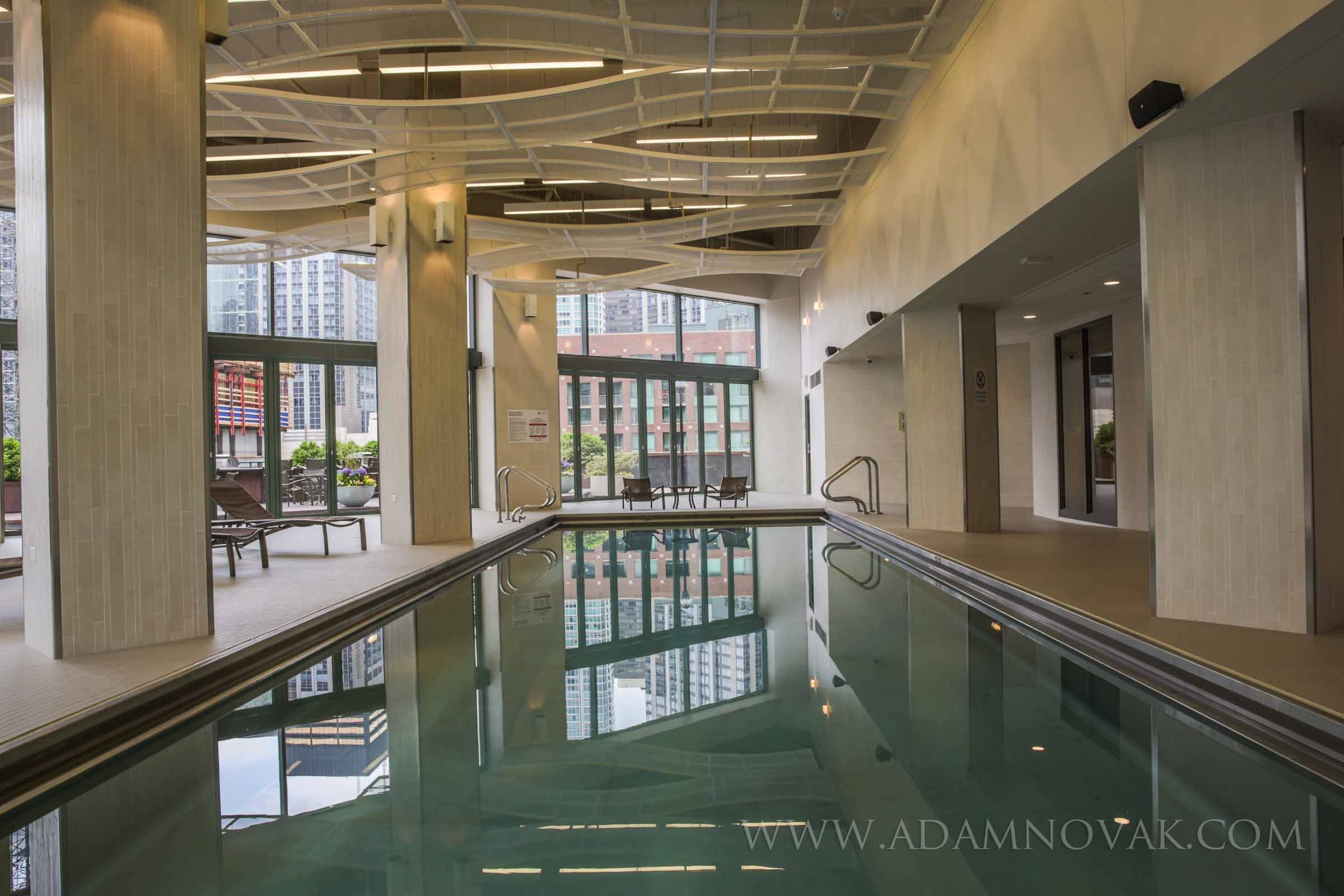 Indoor swimming pool area with large windows, lounge chairs, and modern ceiling design in a high-rise building.