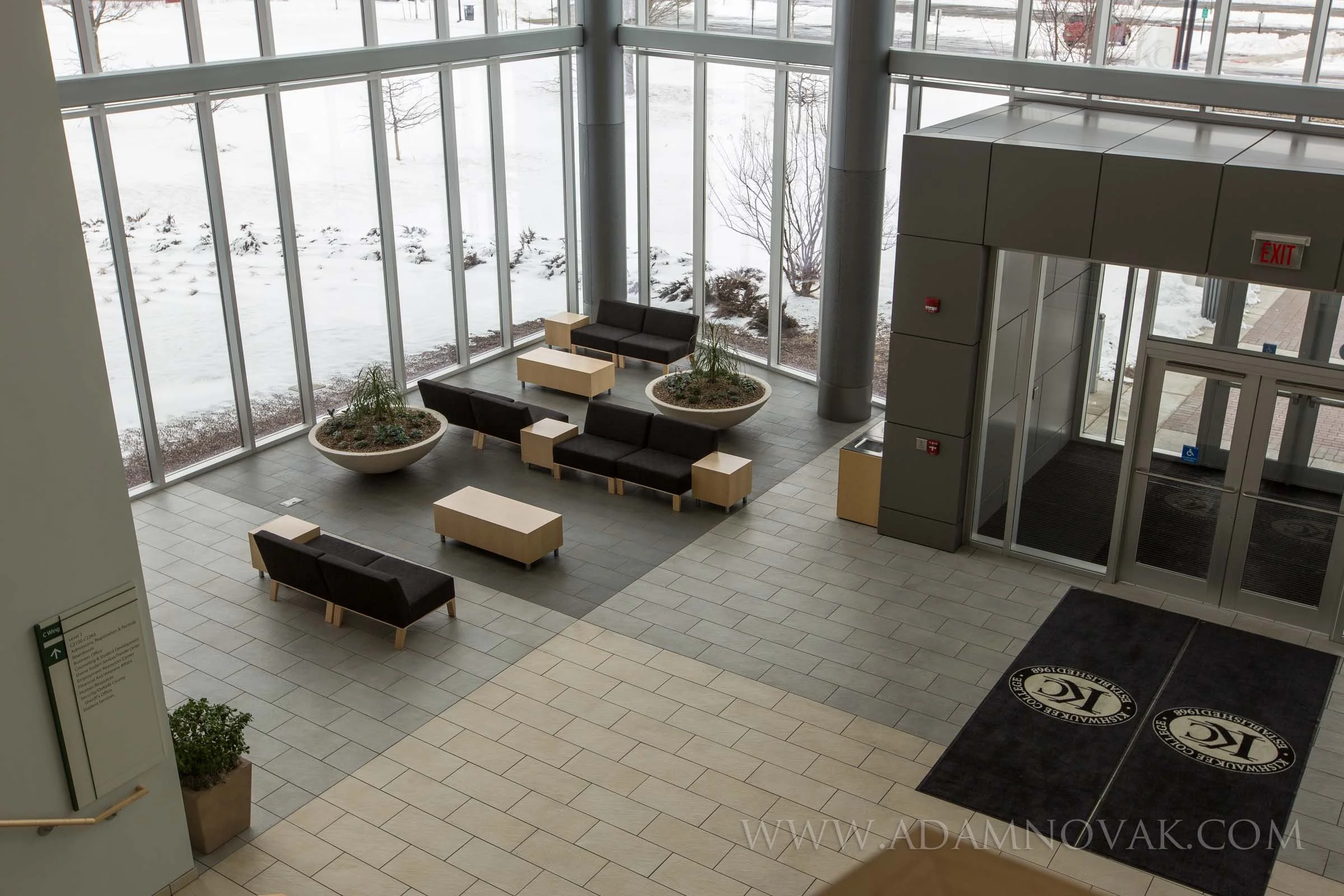 Lobby area with black sofas, beige ottomans, and large planters with plants, surrounded by tall glass windows showing snow outside.