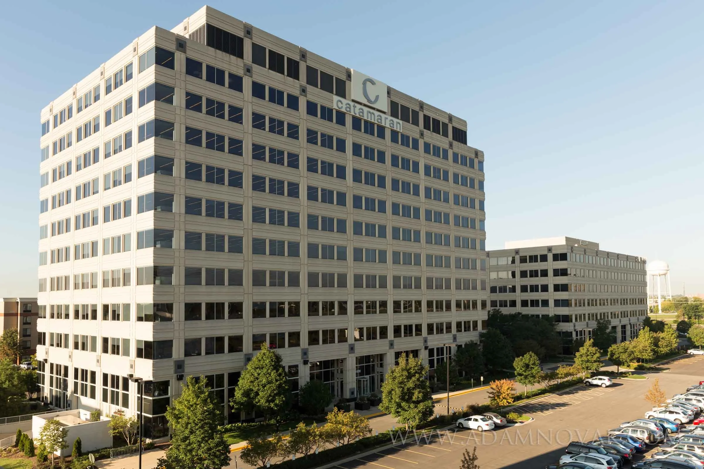 Large modern office building with multiple floors and numerous windows, labeled 'Catamaran,' situated next to a parking lot with cars and surrounded by trees under a clear blue sky.