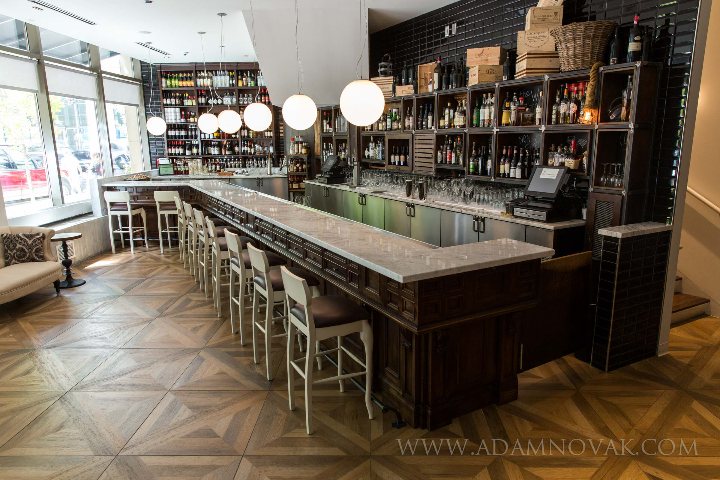 Interior of a modern bar with a marble countertop, white chairs, wine bottles on shelves, and large round hanging lights.