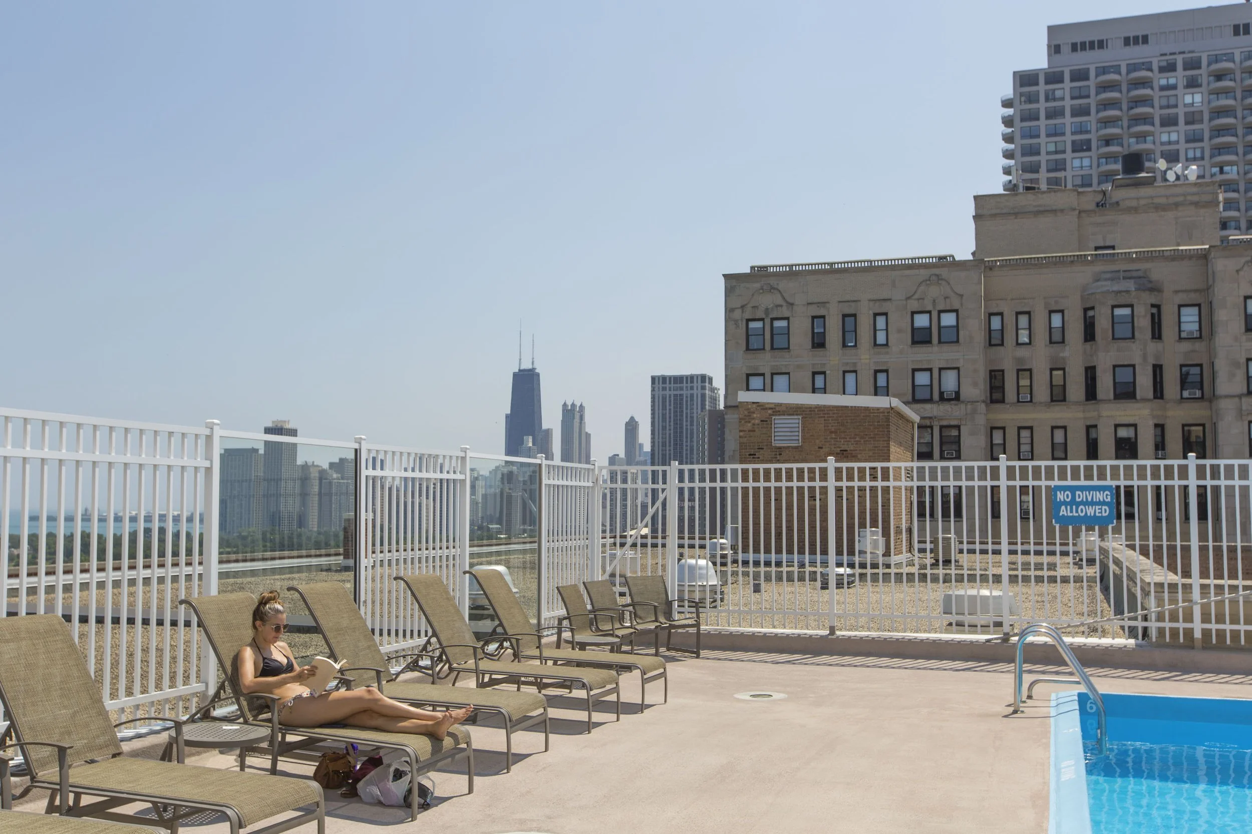 A woman sunbathing on a lounge chair by a rooftop swimming pool, reading a book, with a city skyline featuring tall buildings in the background.