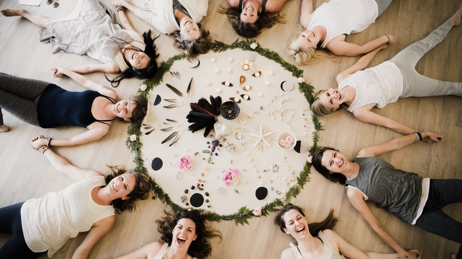 Group of women lying on the floor in a circle around a large, decorated mandala with crystals, feathers, flowers, and candles, smiling and enjoying themselves.