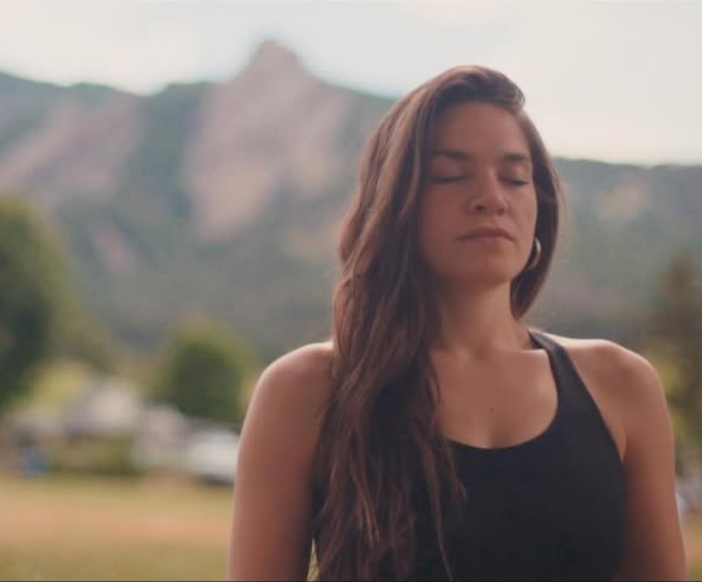 A young woman with long brown hair wearing a black tank top standing outdoors with mountains in the background.