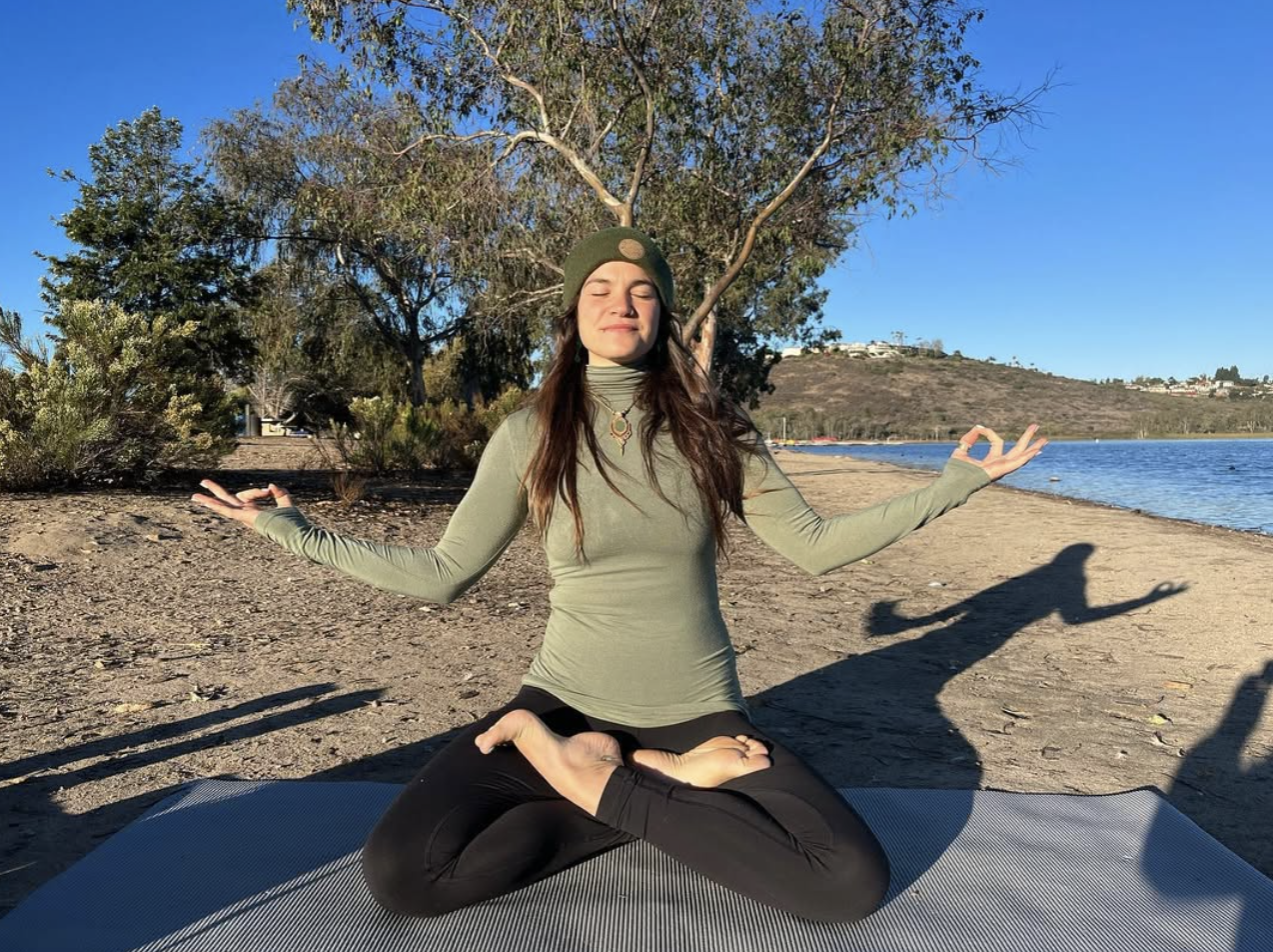 Young woman practicing yoga in a boat pose on a yoga mat by the water with trees and hills in the background.