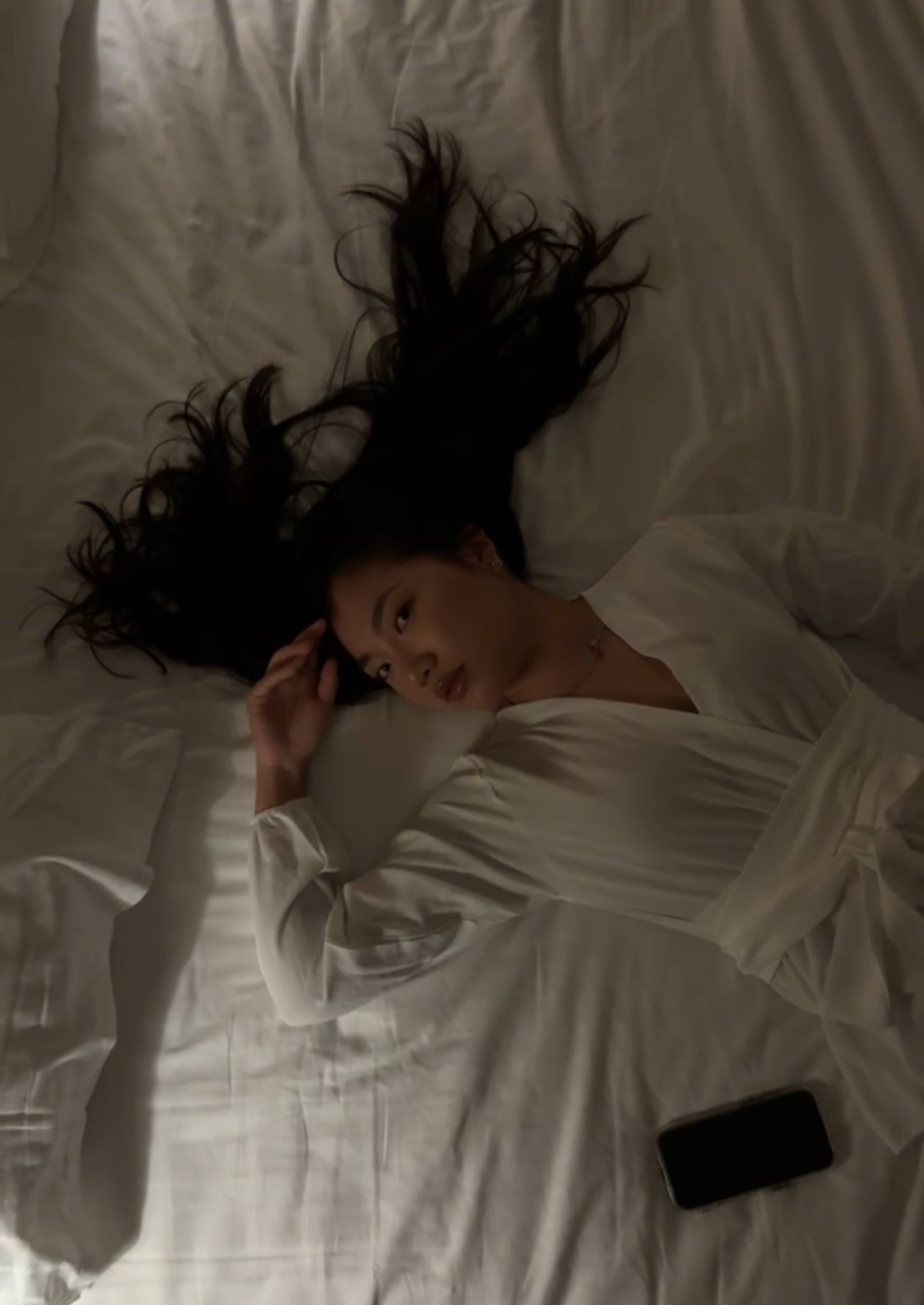 Une jeune femme aux longs cheveux noirs, portant une robe blanche, allongée sur un lit en satin blanc, regardant vers l'objectif. Un téléphone portable se trouve près d'elle sur le lit.