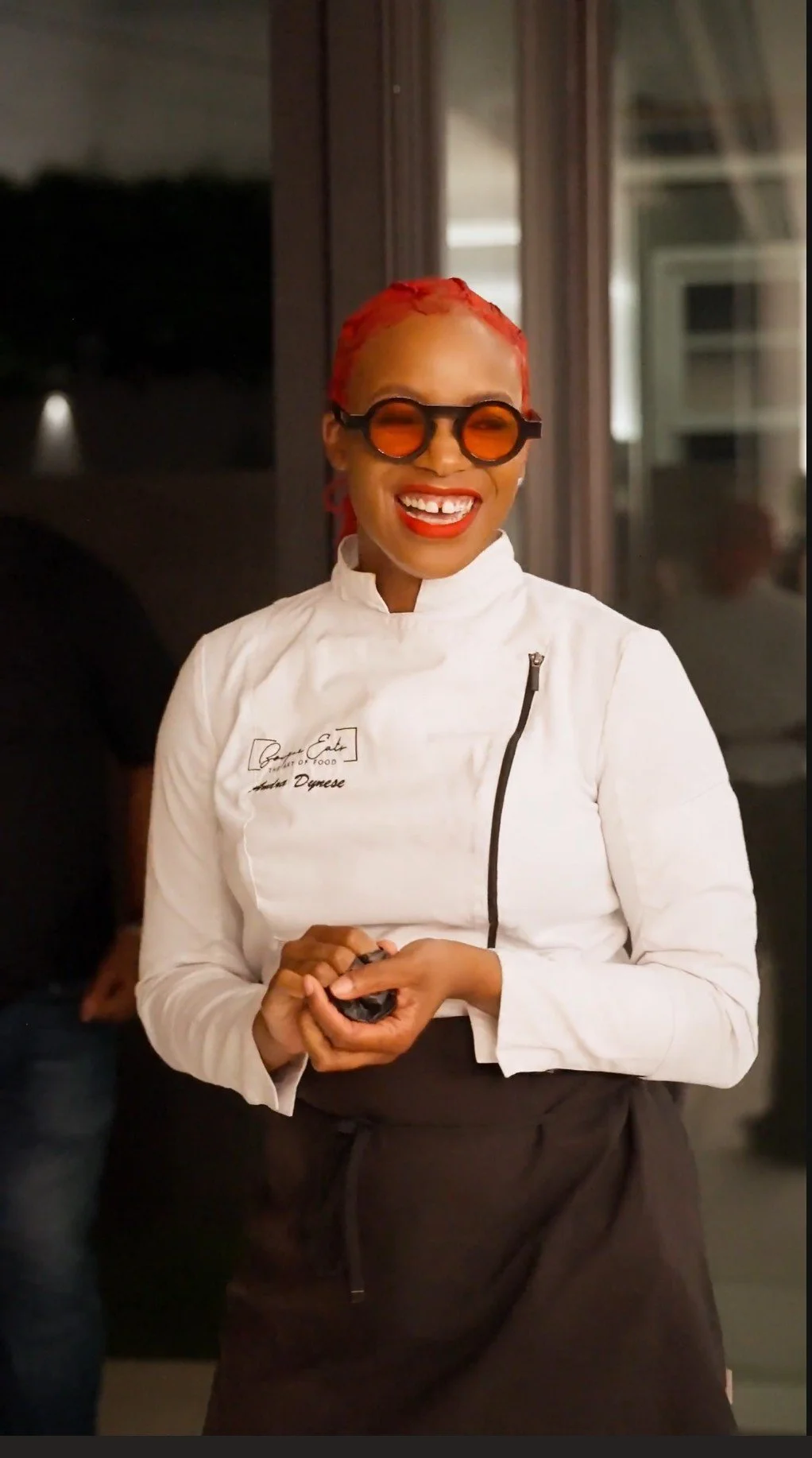 A woman with bright red hair, orange-tinted glasses, and red lipstick, smiling and holding her hands together, wearing a white chef's coat and black apron.
