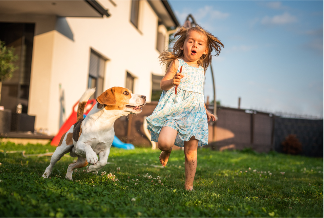 A young girl running barefoot on a grassy backyard with a beagle dog, both appearing excited and energetic.