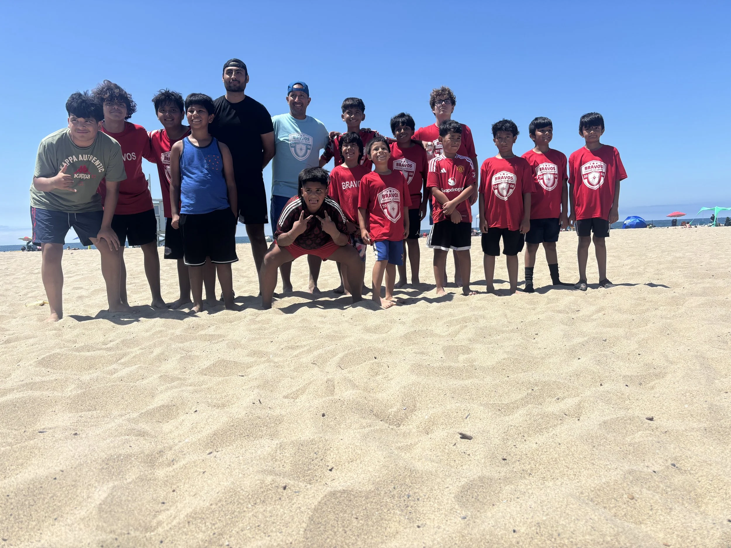 A group of children and two adult men standing on a sandy beach under a bright blue sky with a few colorful umbrellas in the background.