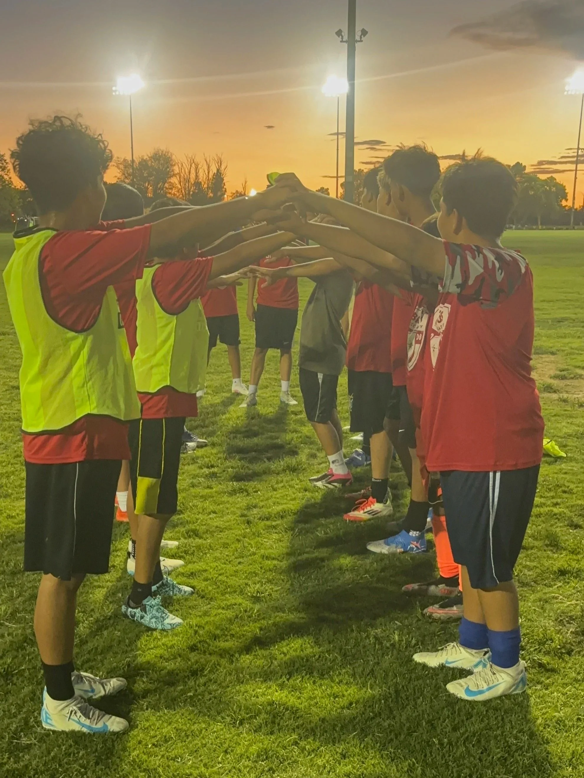 A group of young soccer players standing face to face in two lines, extending their arms and hands towards each other in a team cheer or warm-up on a grassy field during sunset.
