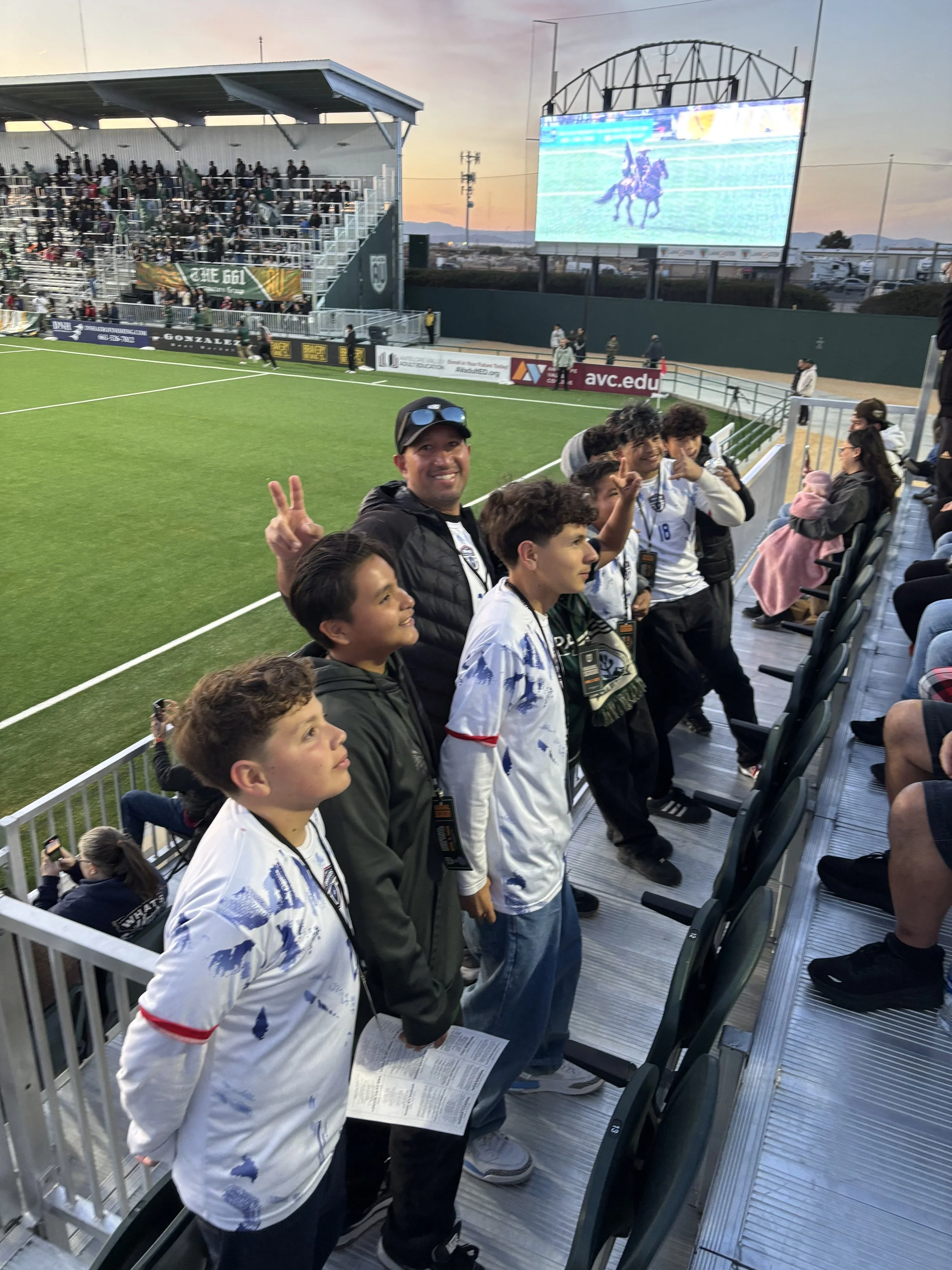 Group of young boys and an adult at a soccer game, standing in a row on the sidelines, with some making peace signs, on a sports field during sunset.