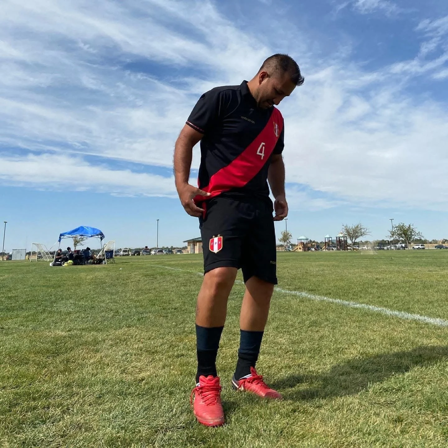Gerardo Bravo standing on a grassy soccer field, wearing a black and red sports uniform with the number 4, adjusting his uniform, with a blue sky and scattered clouds in the background.