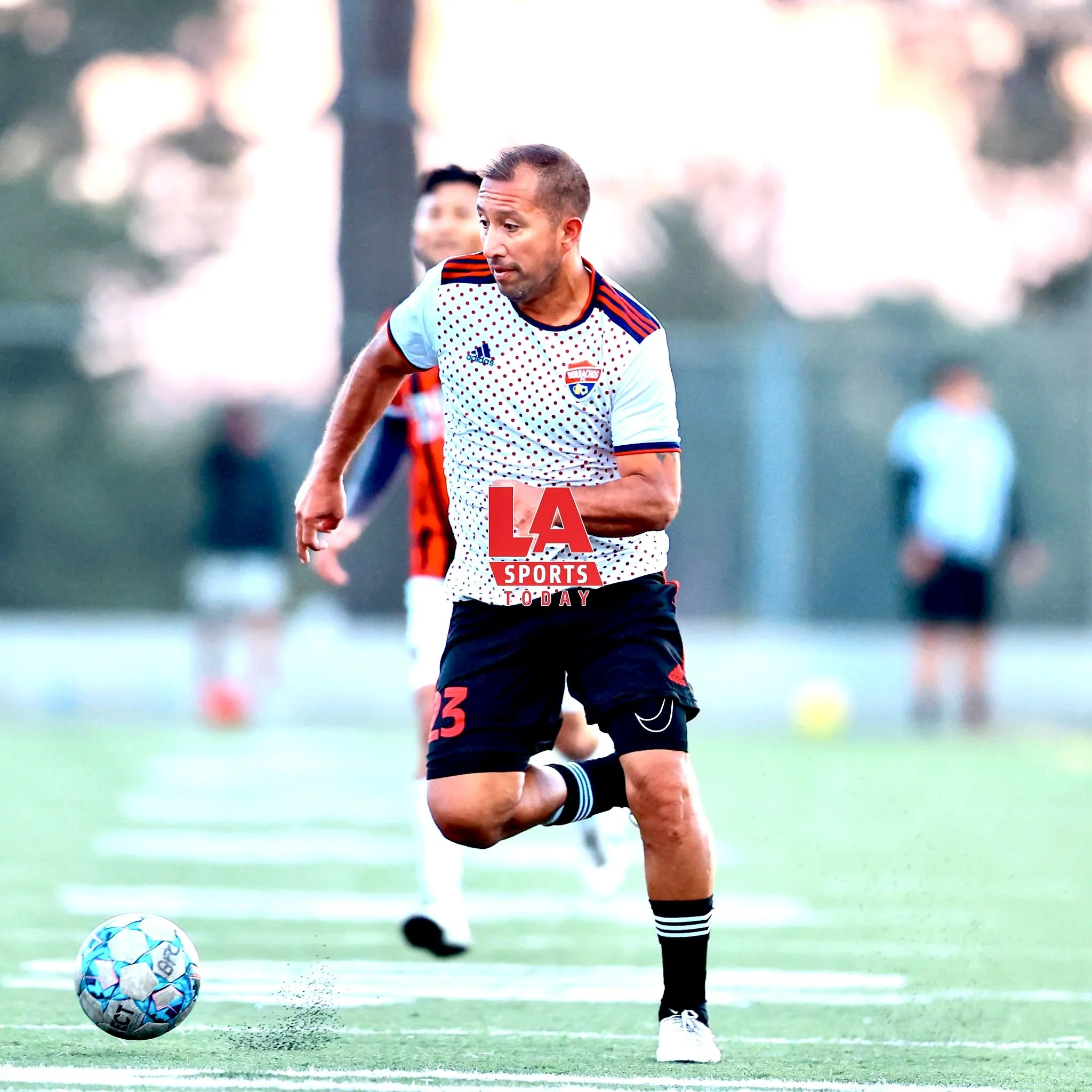 Gerardo Bravo playing soccer on a field during daytime, wearing a white and red jersey, black shorts, and black and white socks, with a soccer ball near his foot.