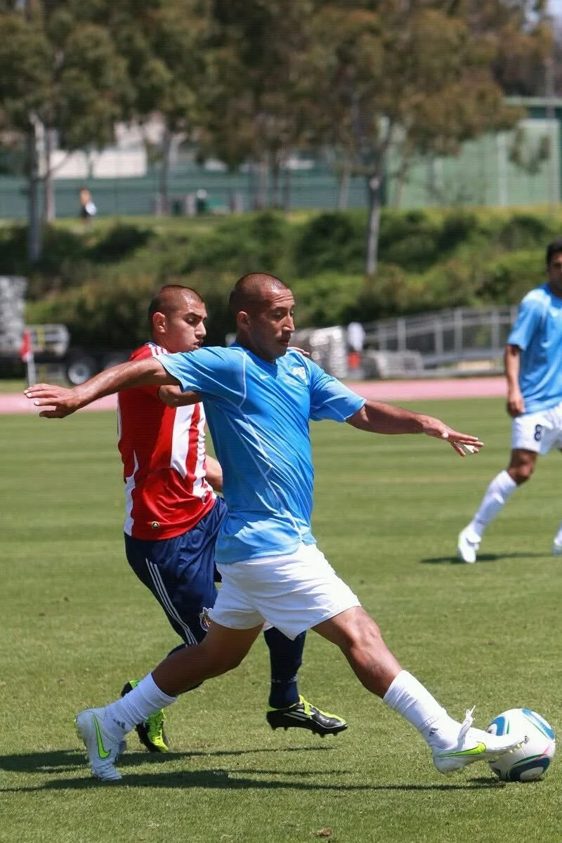 Soccer players competing on the field, with one player in a blue jersey about to kick the ball while another player in a red jersey tries to block him.