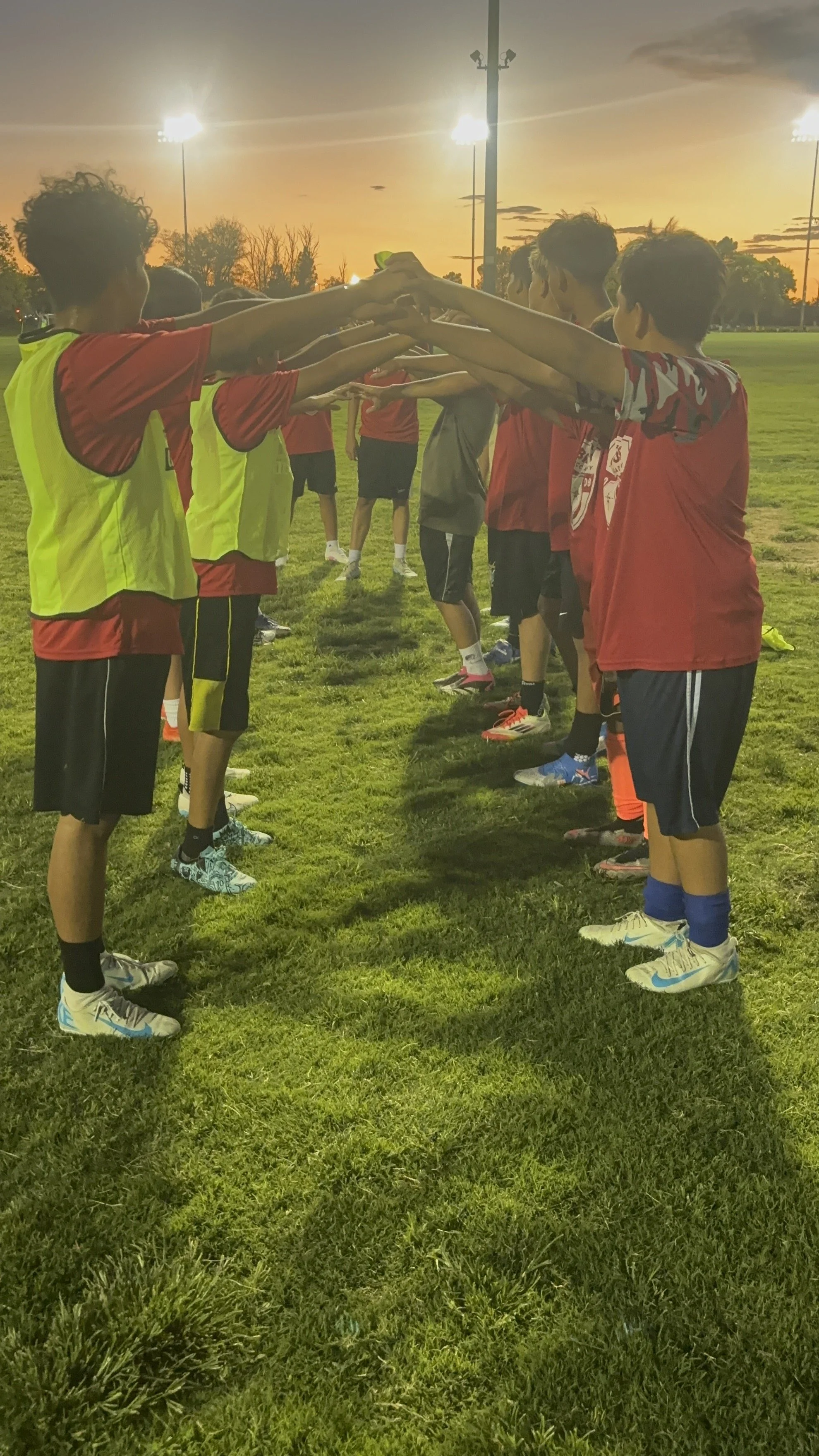 A group of young soccer players standing face to face in two lines, extending their arms and hands towards each other in a team cheer or warm-up on a grassy field during sunset.