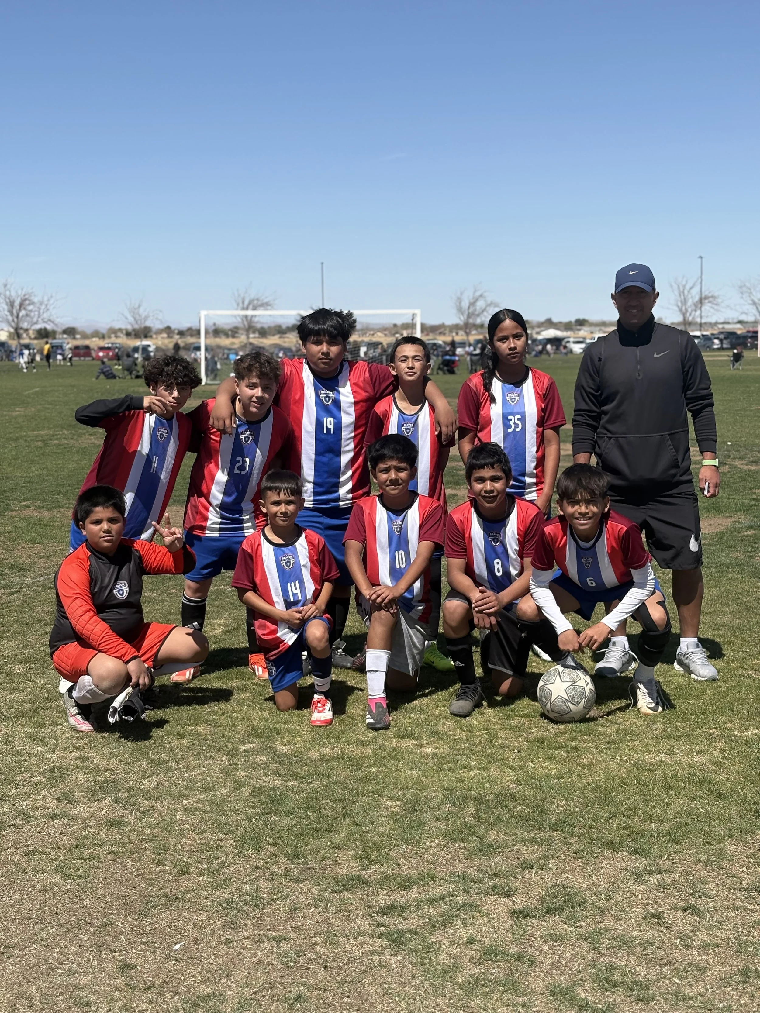 BSA UNITED in red, white, and blue uniforms poses with their coach on a soccer field. The team is standing and kneeling around a soccer ball, with a goal in the background and a parking lot with cars behind the field under a clear blue sky.