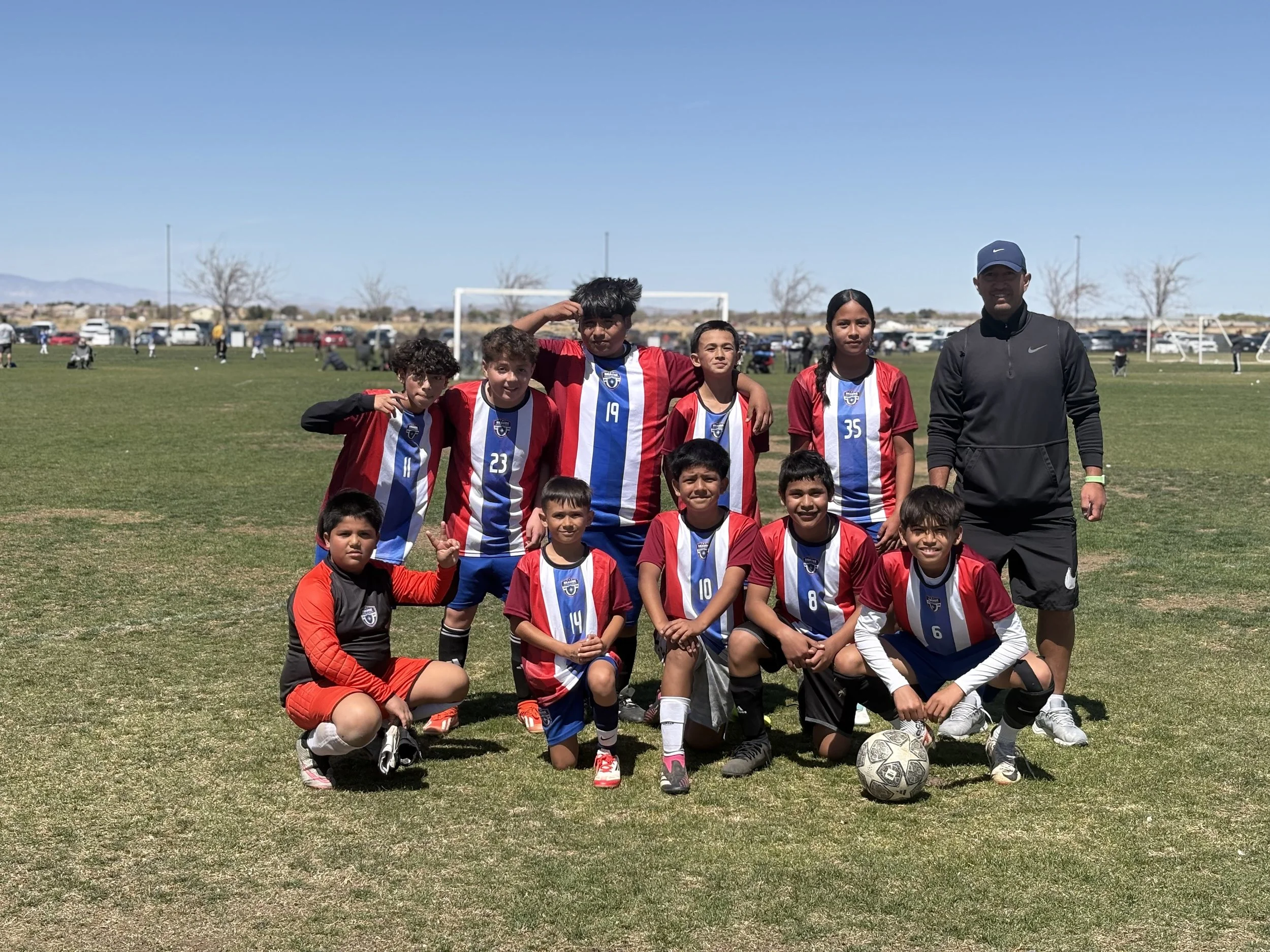 A youth soccer team, wearing red, white, and blue uniforms, posing for a photo on a grassy field with their coach. The team includes children with different jersey numbers. Some children are standing, some are kneeling, and there's a soccer ball in f