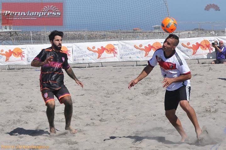 Two men are playing beach soccer on a sandy field, with one man about to head the orange soccer ball while the other man looks on. In the background, there are banners and a person taking photographs.
