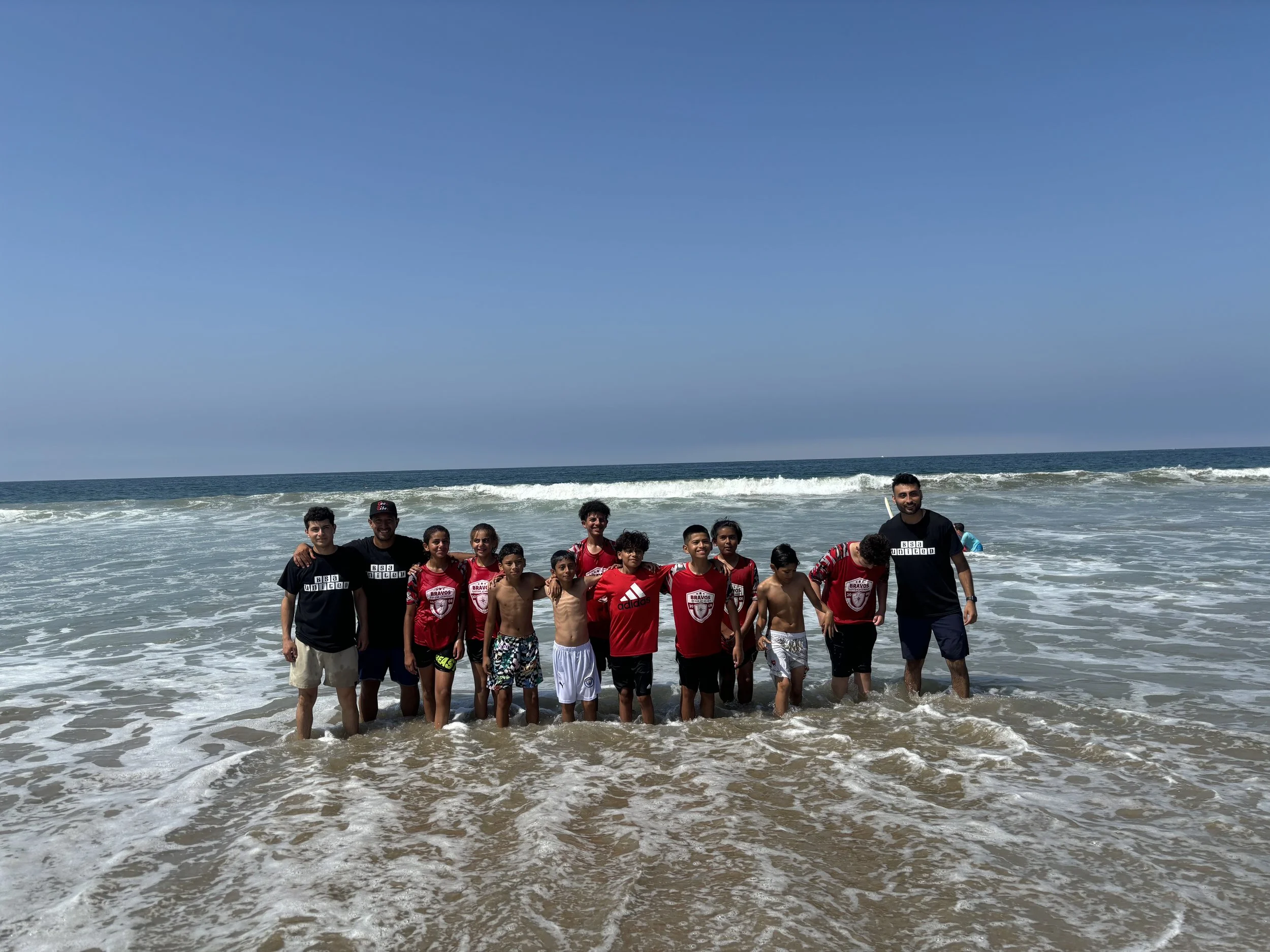 Group of children and adults standing in the ocean on a sunny day, some wearing red sports jerseys and others casual clothes, with waves and clear blue sky in the background.