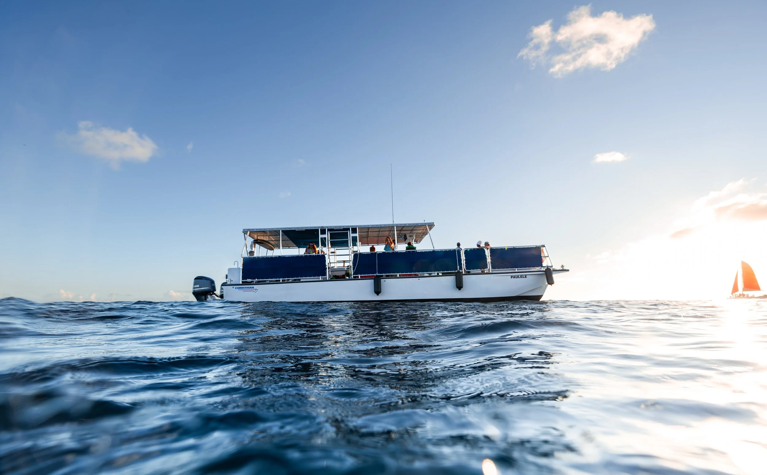 A white and blue boat floating on the ocean with a clear sky and a sailboat in the distance at sunset.
