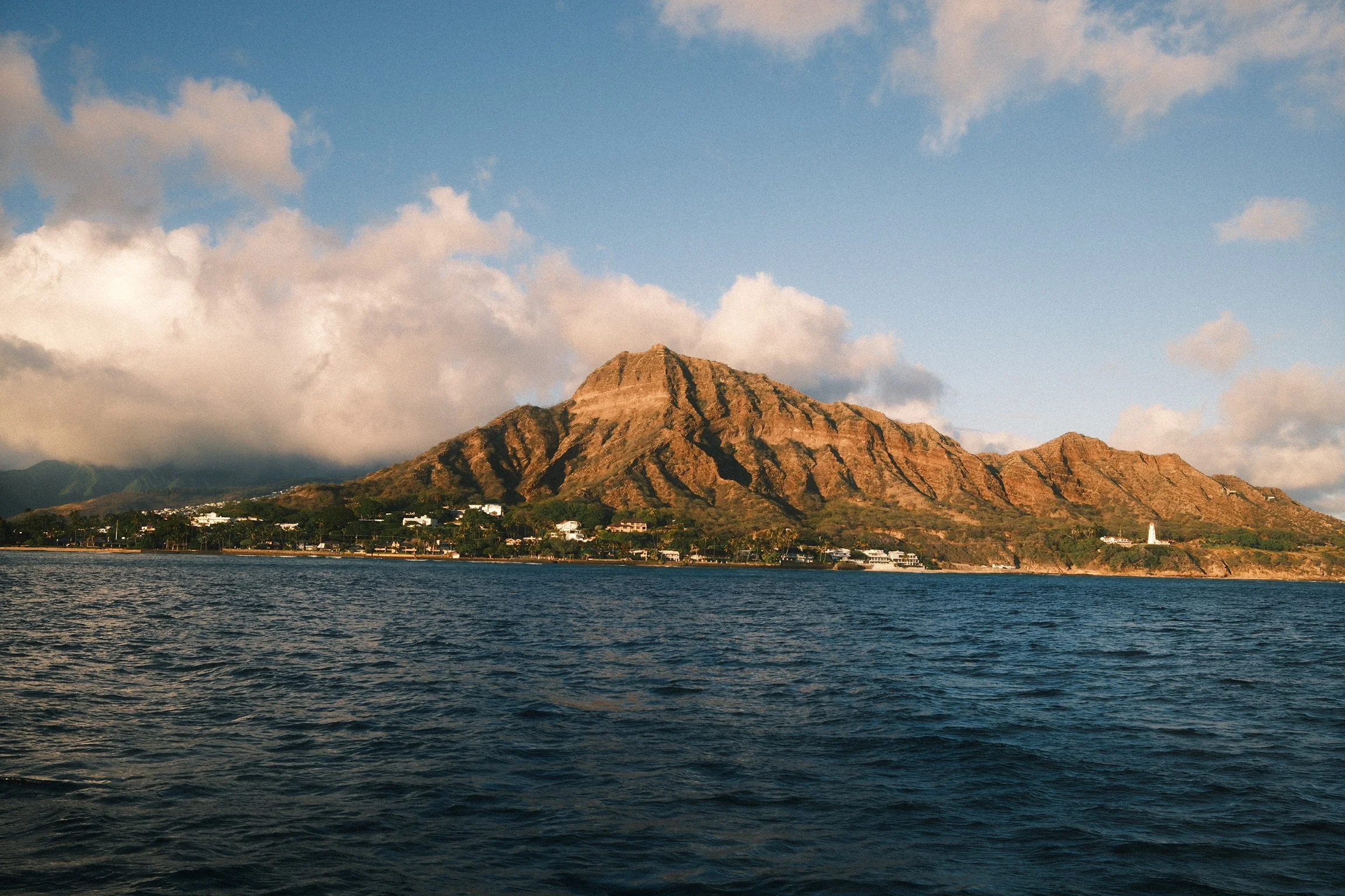 A mountain with a flat top and steep sides, likely a volcanic formation, with a shoreline and houses at its base, under a partly cloudy sky.